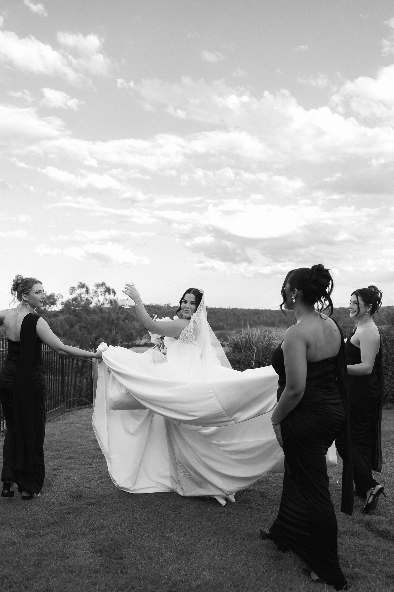 Bridesmaids in black gowns help bride with flowing white dress in black and white photo.