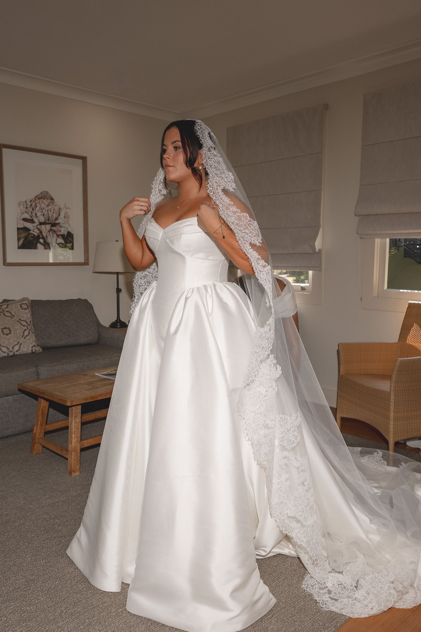 Elegant bride in strapless satin ball gown with lace cathedral veil poses indoors before wedding ceremony.