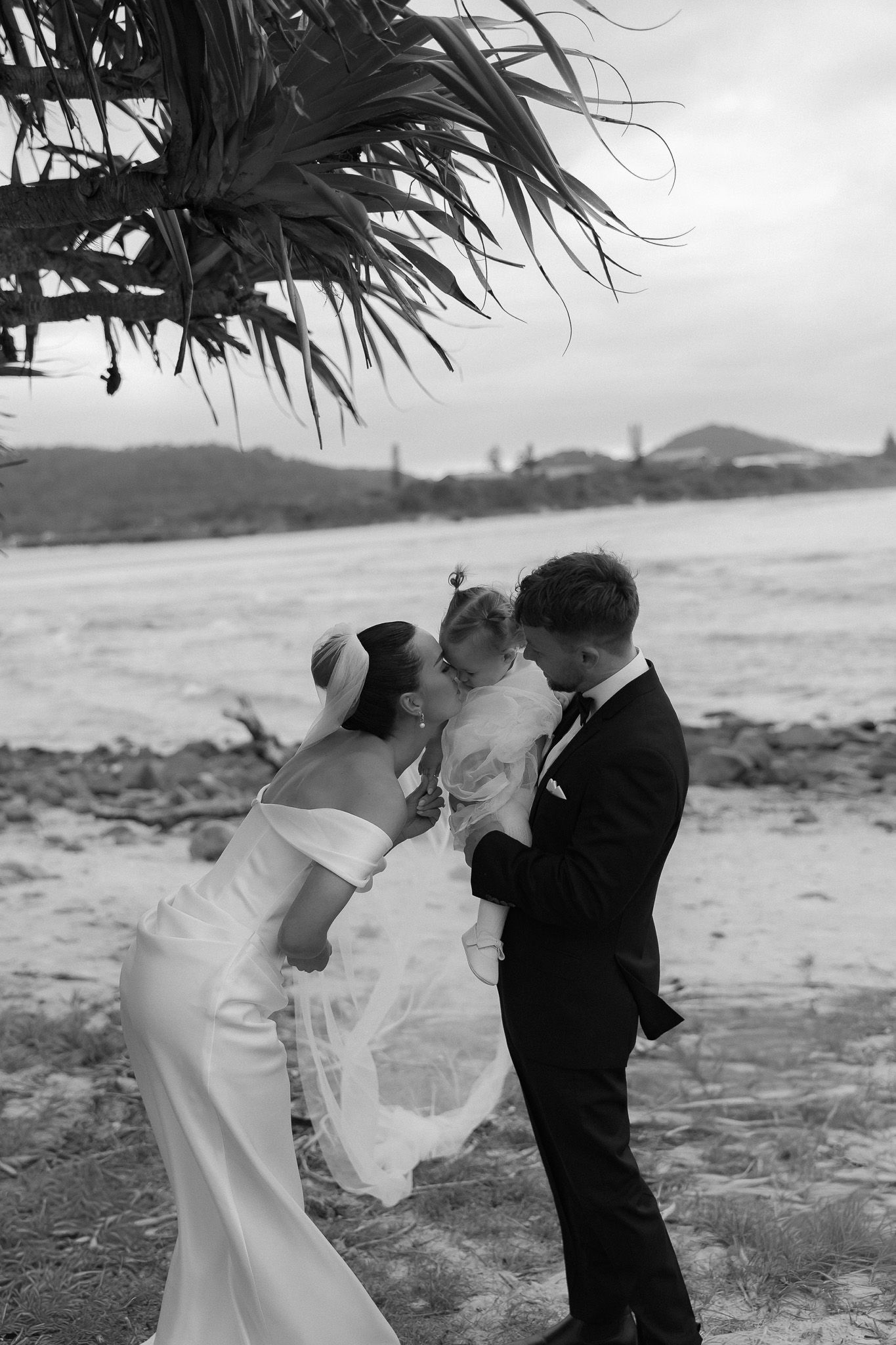 Black and white beach wedding photo of bride and groom kissing their child under a palm tree with ocean backdrop.