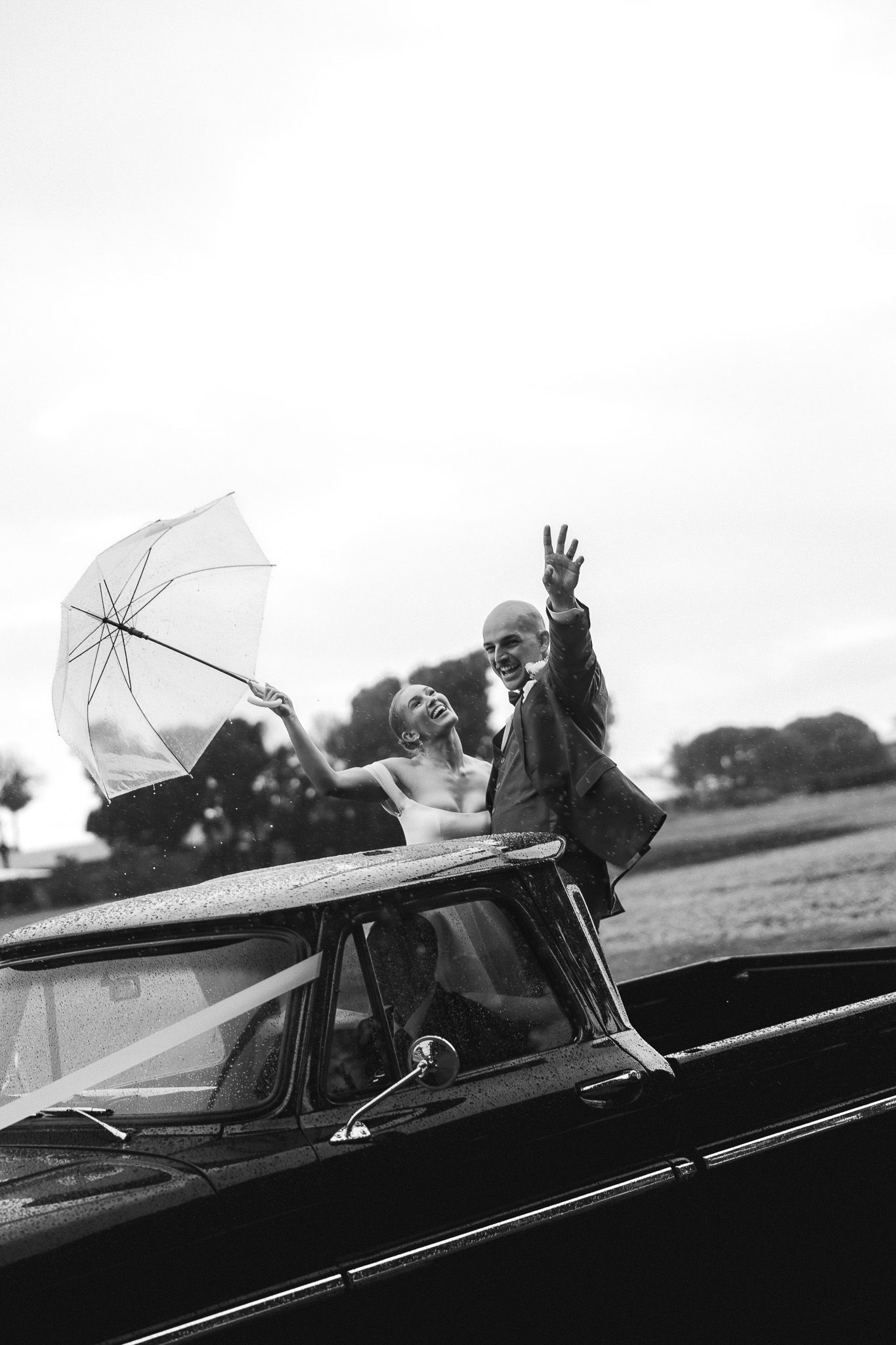 Black and white photo of joyful newlyweds waving from vintage convertible car with umbrella.