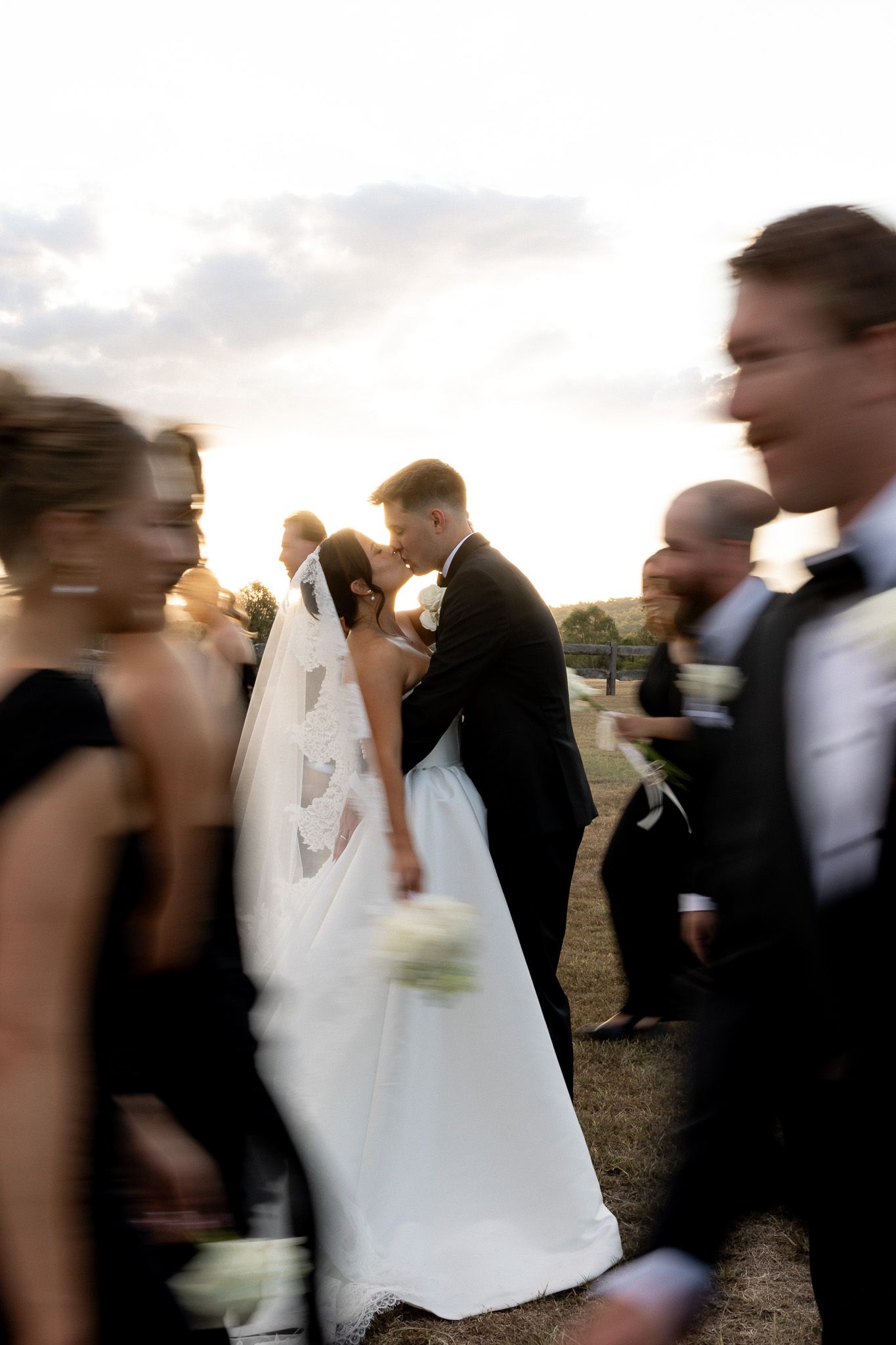 Bride and groom share a kiss surrounded by blurred wedding party in motion at sunset.