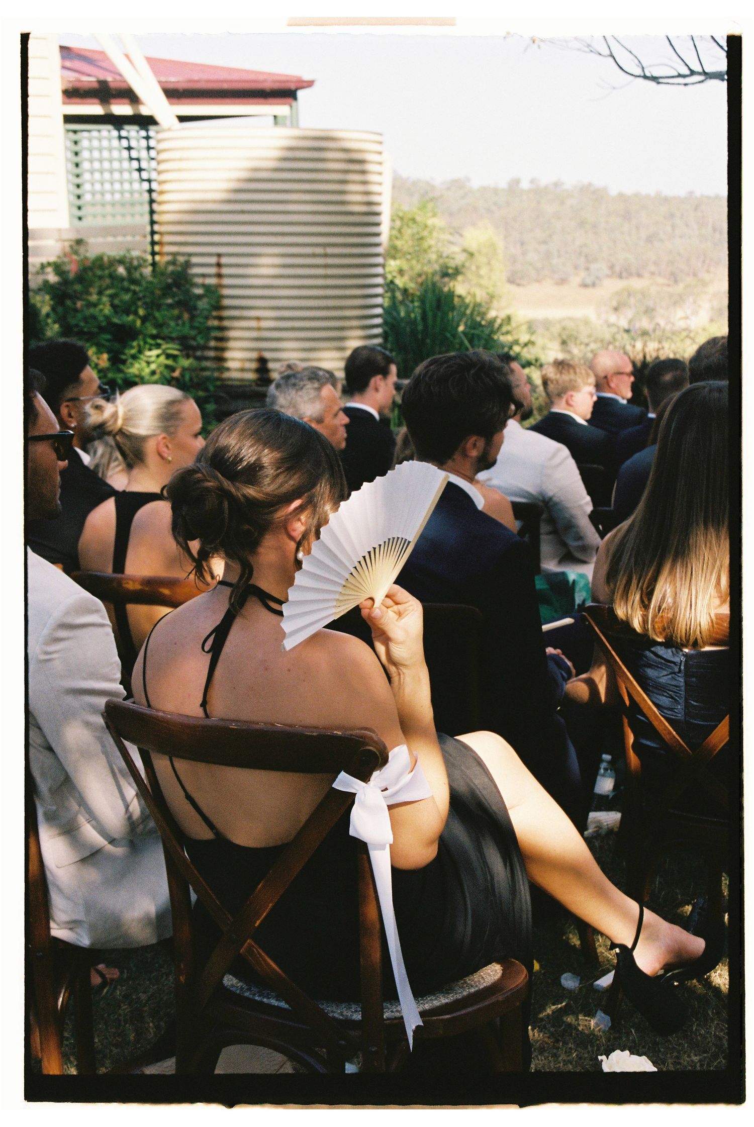 Wedding guest holding a white hand fan at an outdoor ceremony on a warm sunny day.