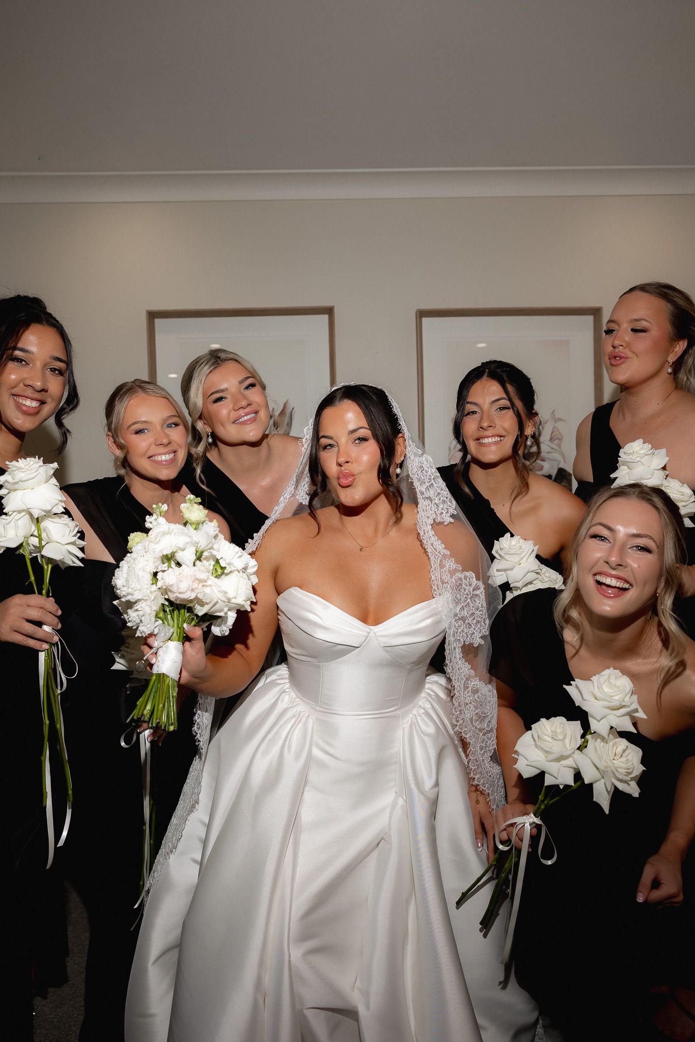 Bride in white strapless gown posing with bridesmaids in black dresses holding white bouquets.