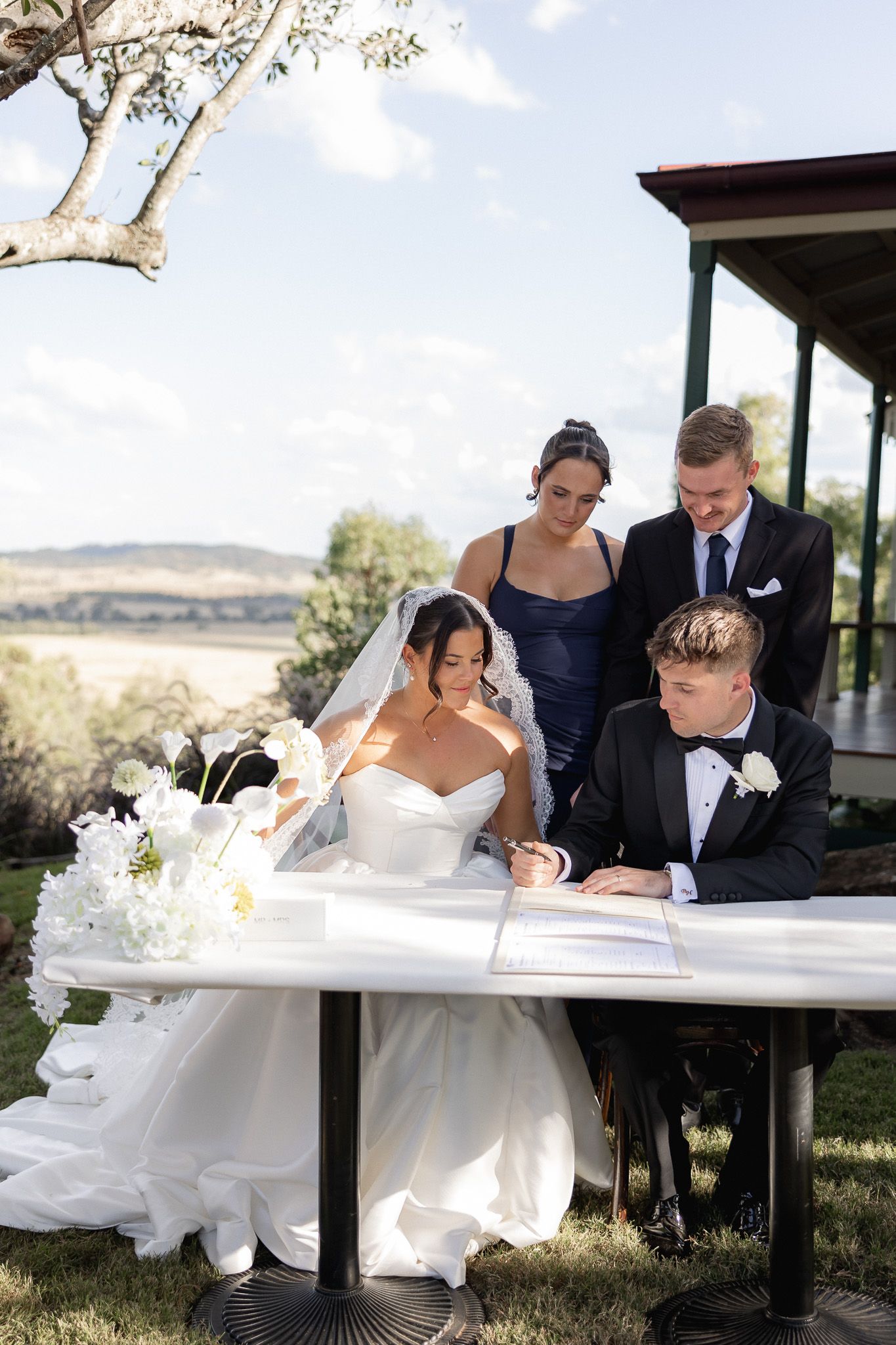 Bride and groom signing wedding register outdoors at a table with white floral arrangements, witnessed by bridesmaids.