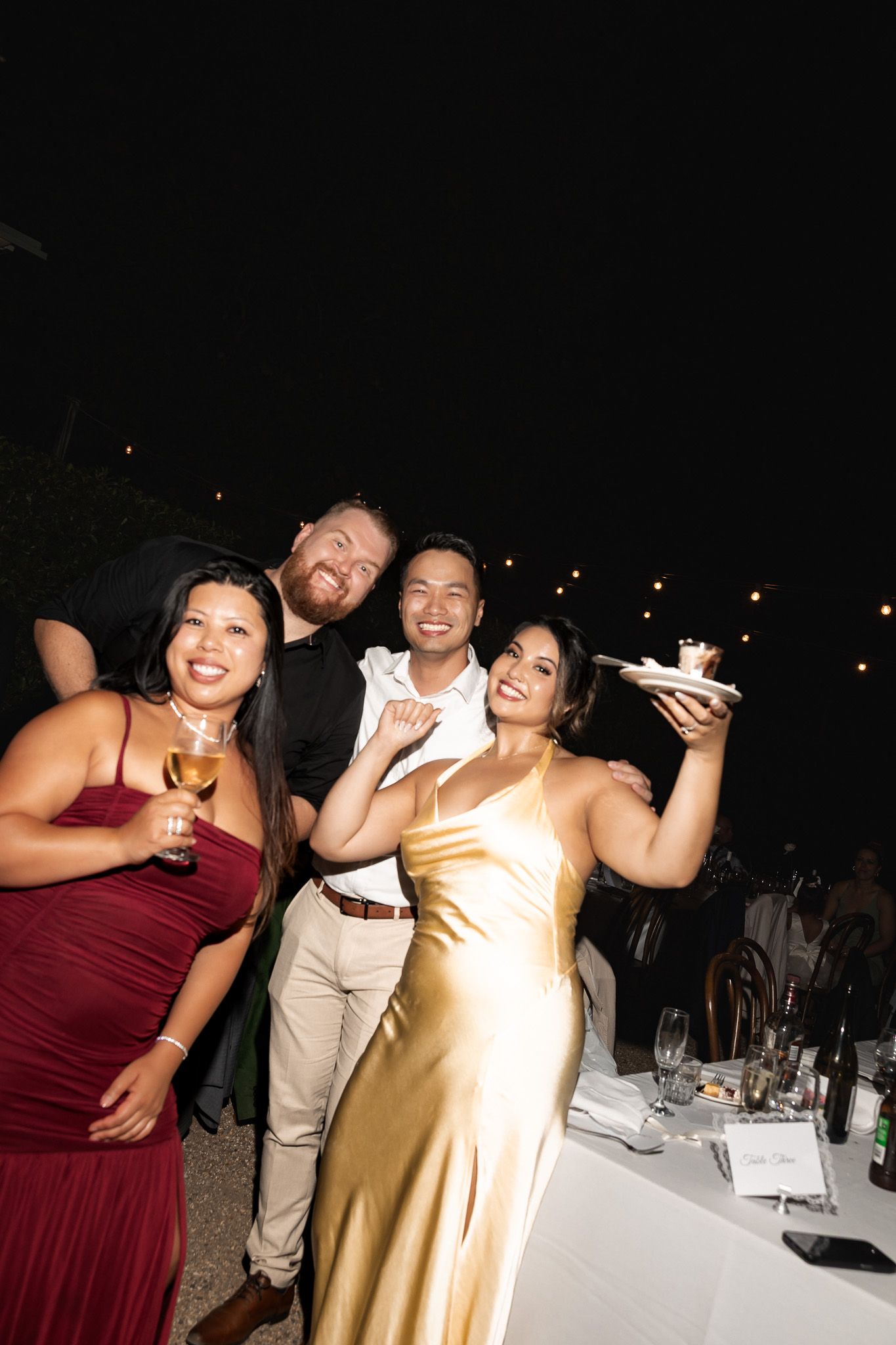 Four wedding guests in formal attire smiling and posing with cake at outdoor evening reception.