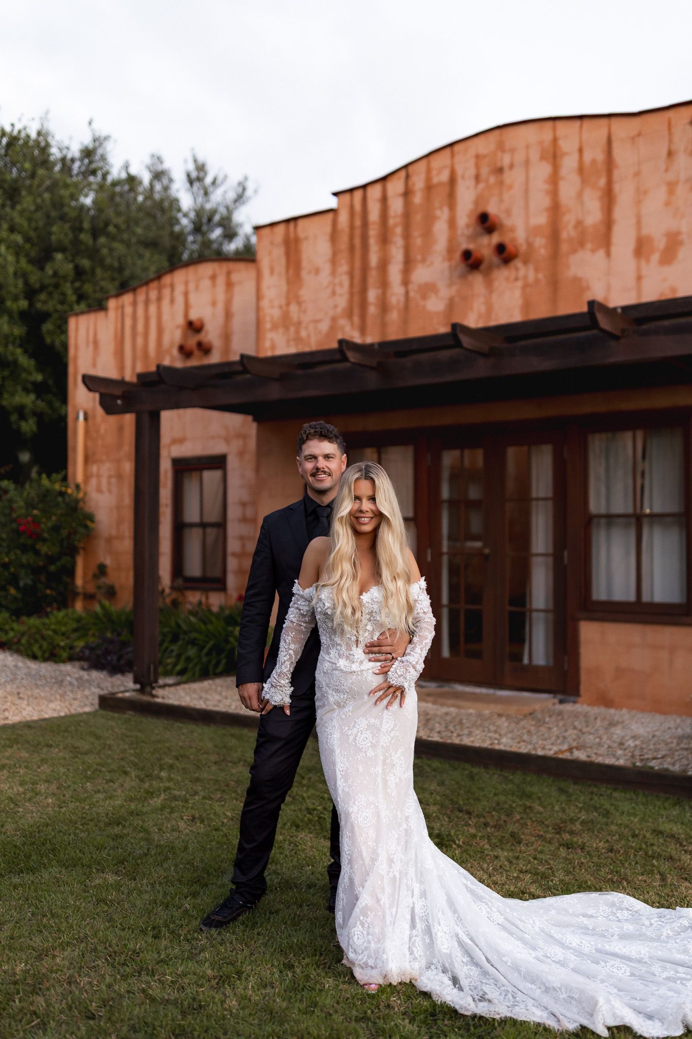 Newlywed couple posing in front of rustic terracotta building, bride in lace gown, groom in black suit.