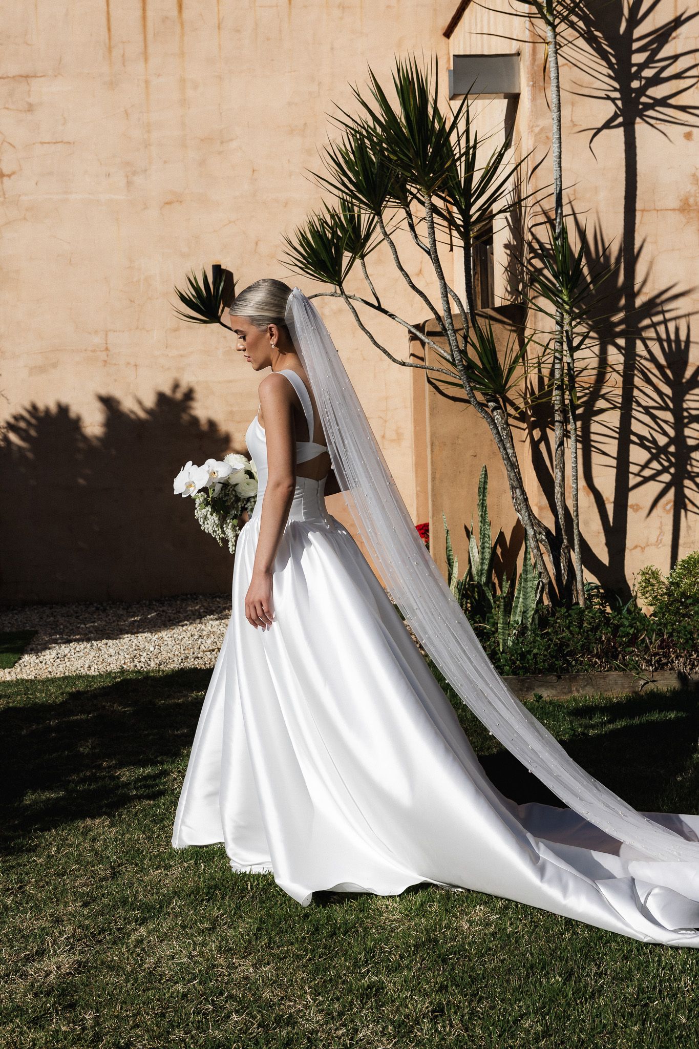 Bride in white ball gown and long veil holds bouquet outdoors against a warm stone wall.