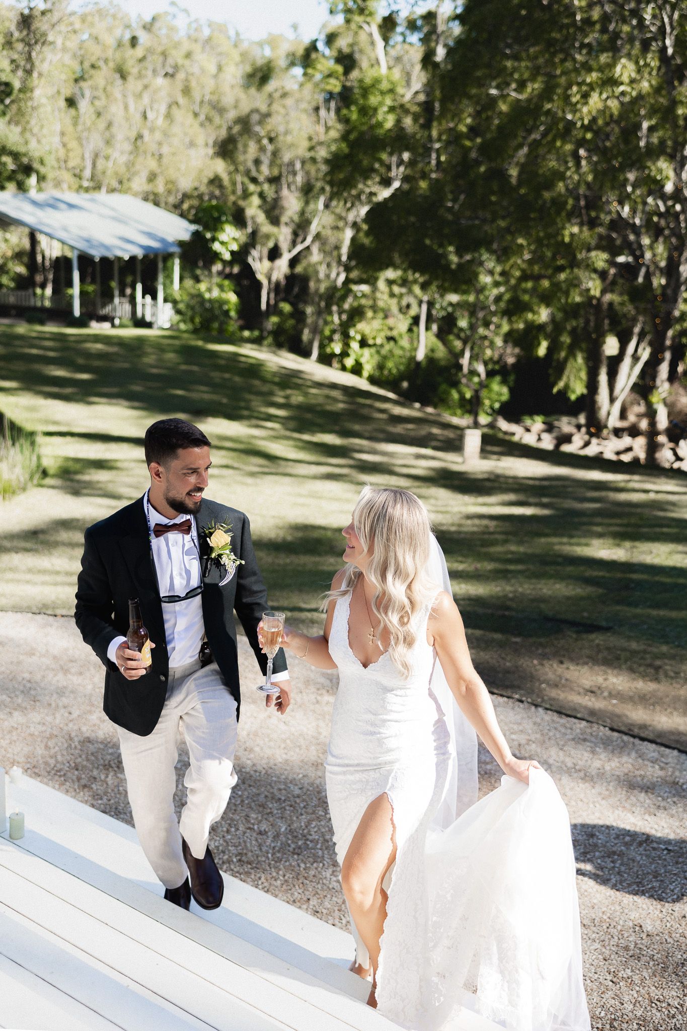 A smiling bride and groom walking with champagne glasses through a lush green outdoor venue.