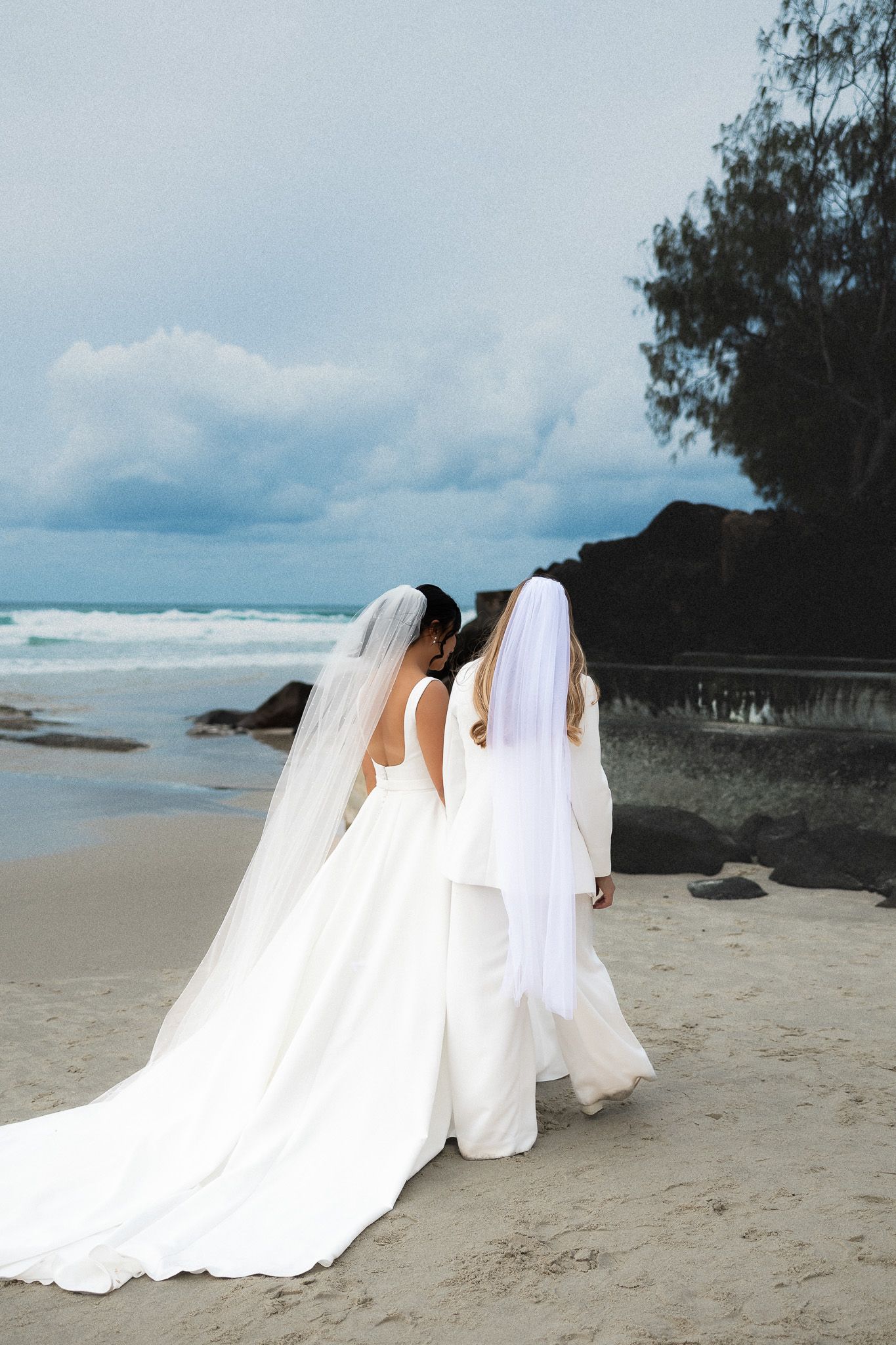 Two brides in white wedding gowns and veils walking along a beach together.