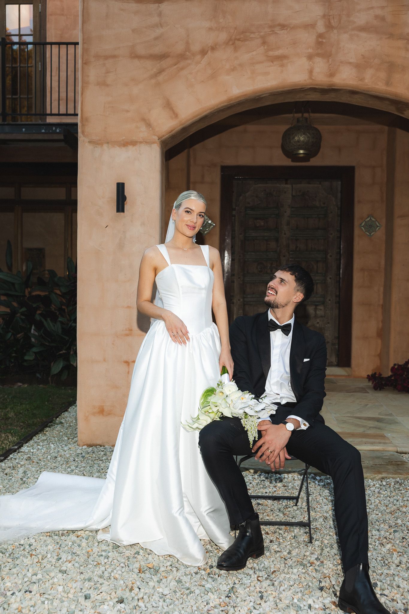 Bride in white satin gown holds bouquet beside groom in black tuxedo seated outside elegant venue.