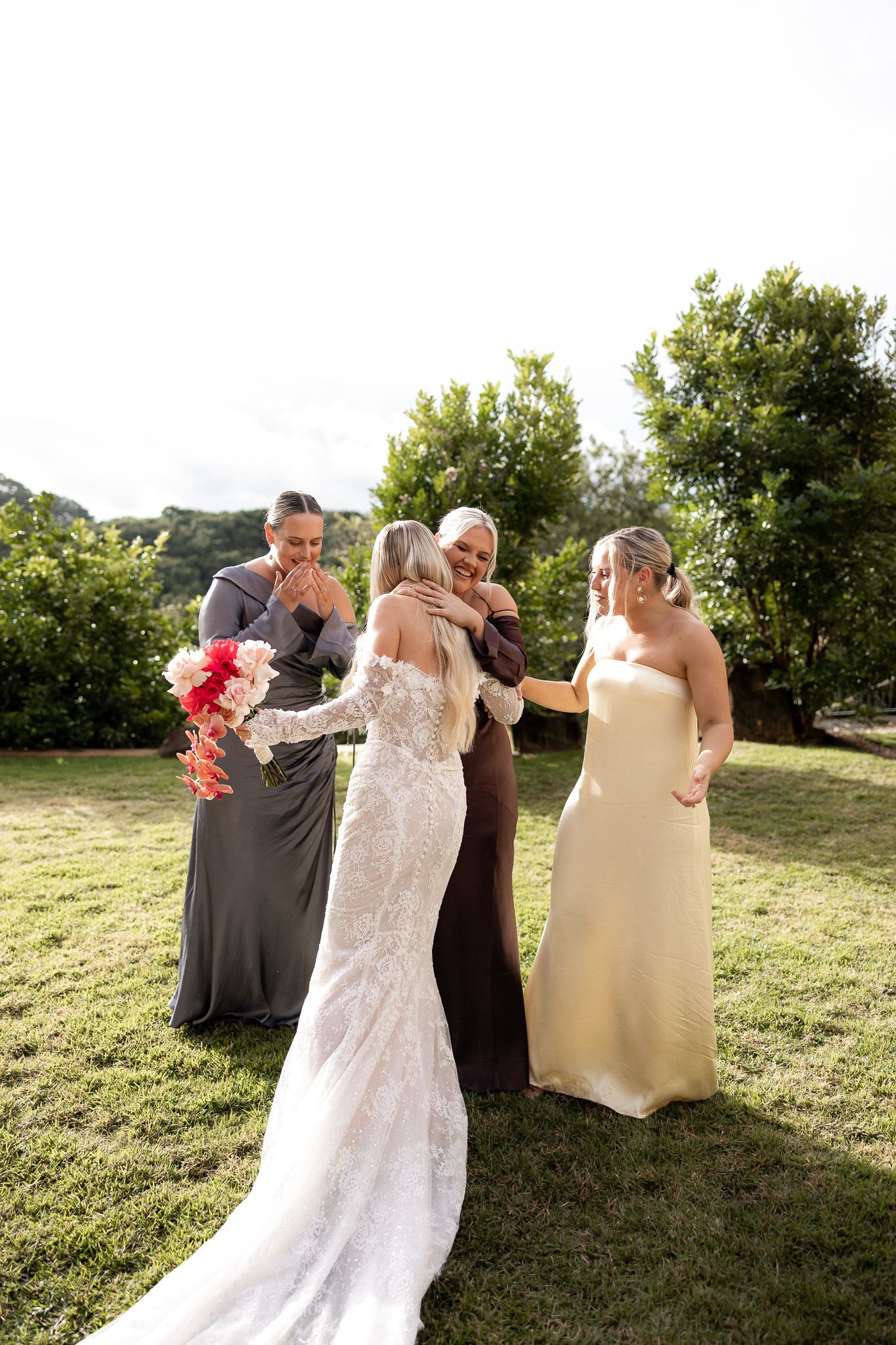 Bride in lace gown hugging guest outdoors at wedding, with bridesmaids in gray and gold dresses holding pink bouquet.