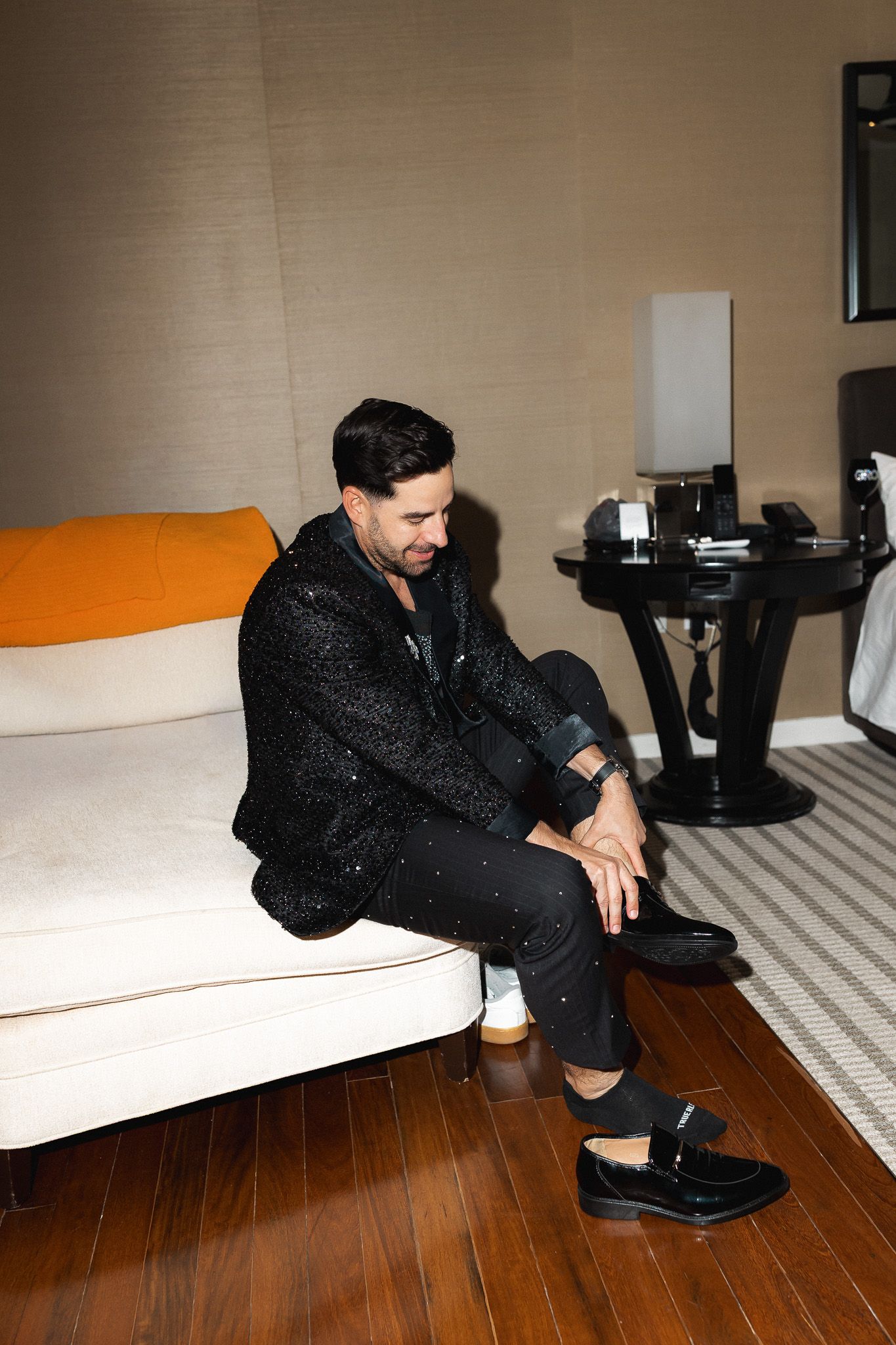 Man in black sequin jacket sits on white sofa in elegant hotel room with hardwood floors.