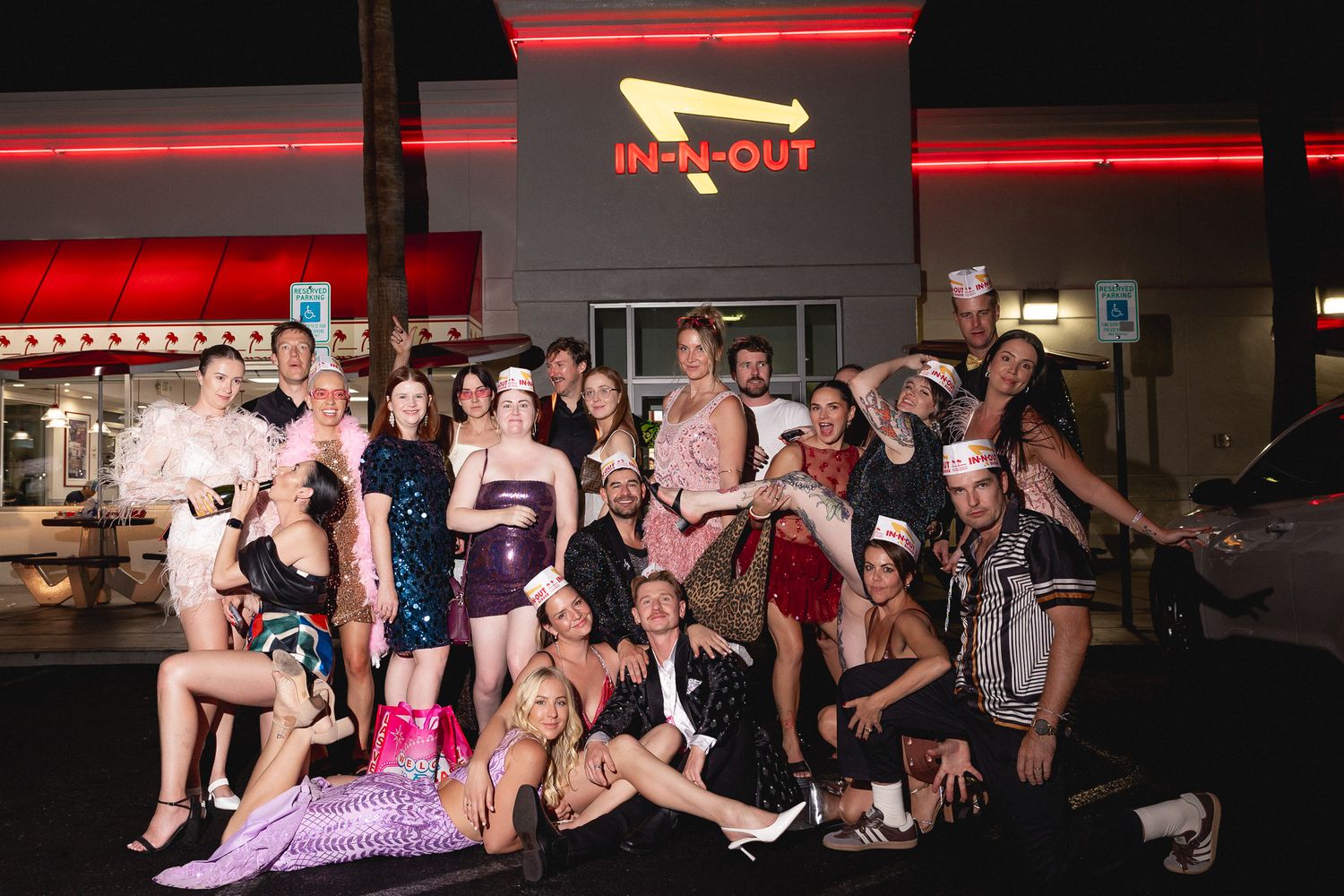 Large festive group poses in glamorous outfits outside an In-N-Out Burger restaurant at night.
