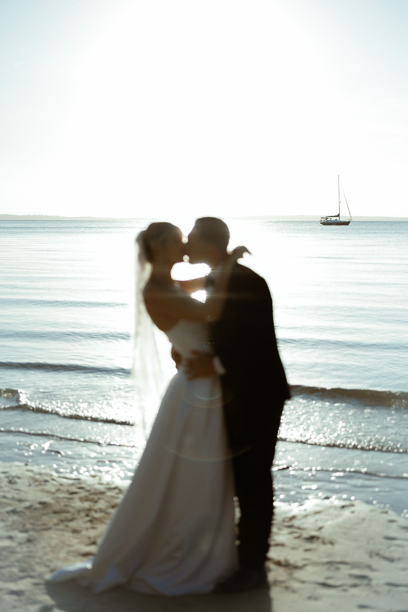 Bride and groom kissing on a sandy beach at sunset with a sailboat in the background.