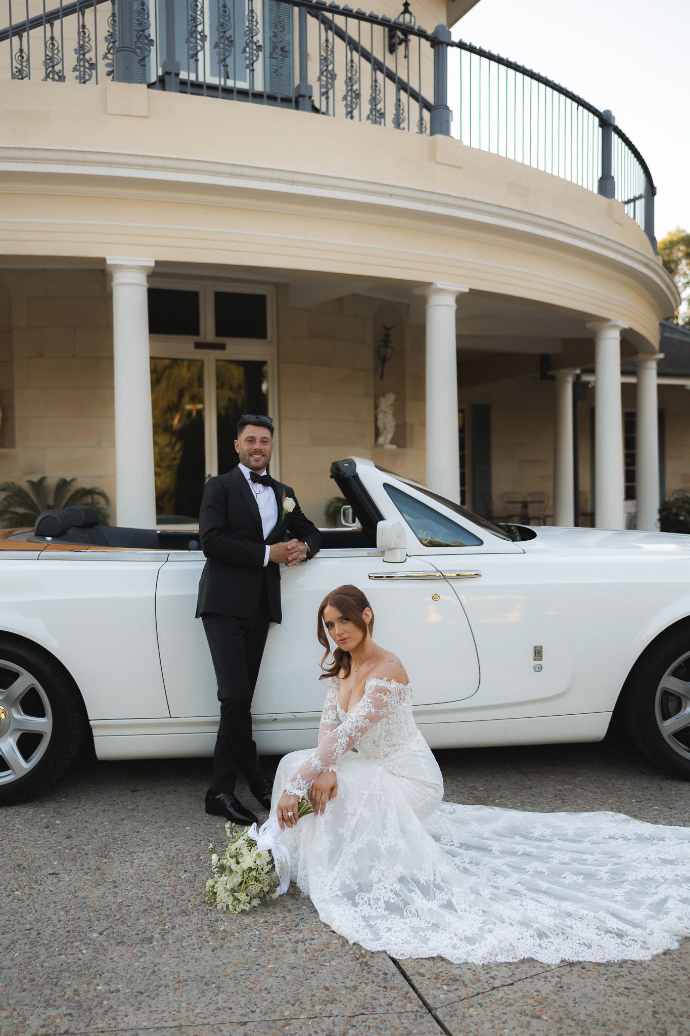 Bride in lace gown sits beside white Rolls-Royce convertible as groom in tuxedo stands behind at elegant venue.