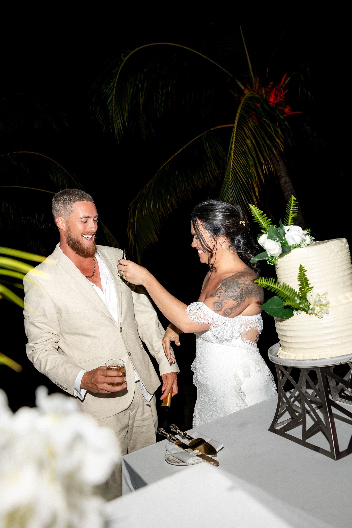 Bride feeds wedding cake to laughing groom during outdoor nighttime tropical wedding reception.