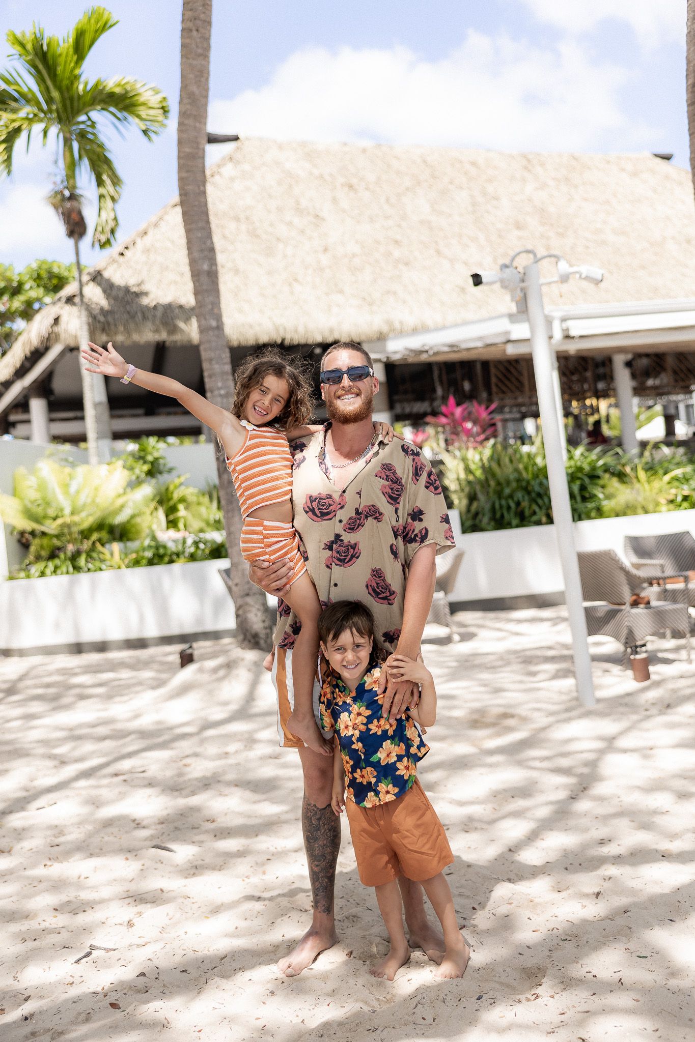 Smiling man in floral shirt holding two children on a tropical resort beach with palm trees.