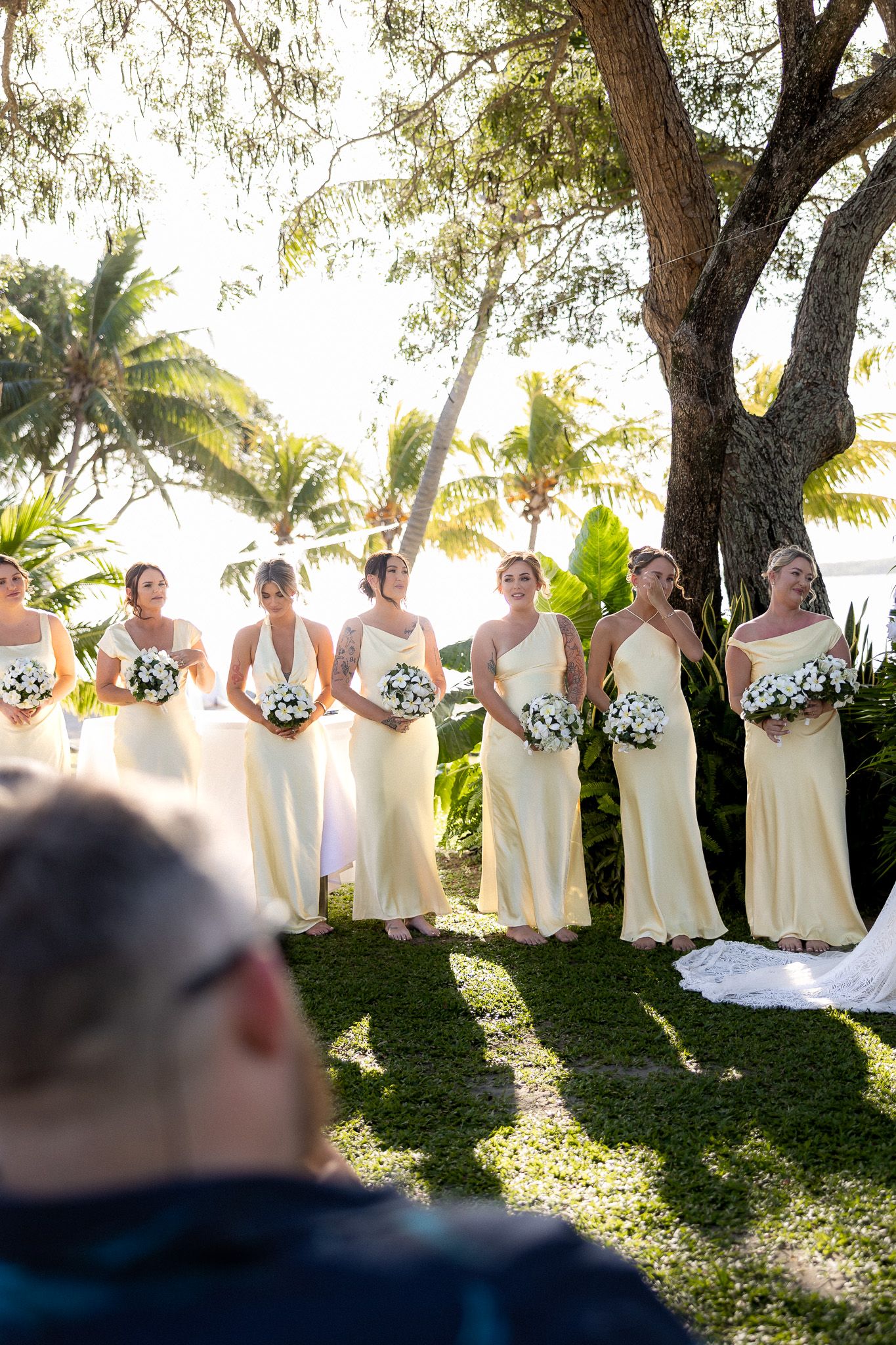 Bridesmaids in yellow dresses holding white bouquets standing in row at tropical outdoor wedding ceremony.