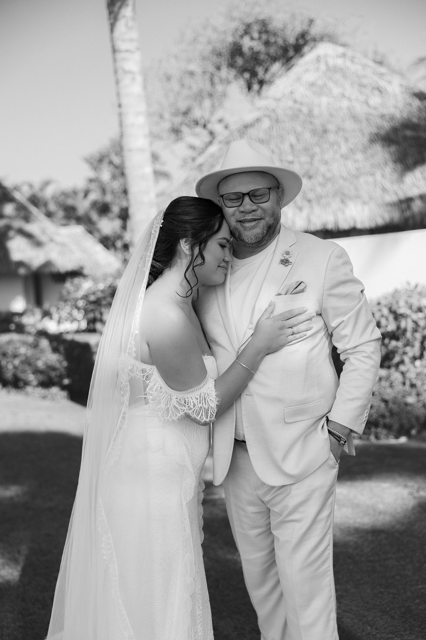 Black and white portrait of smiling groom in panama hat hugging bride in lace wedding dress outdoors.