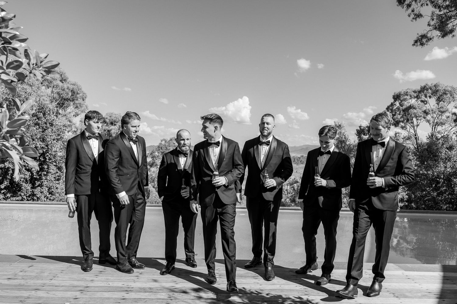 Groom and groomsmen in black tuxedos and bow ties posing outdoors on a rooftop terrace in a black and white photo.