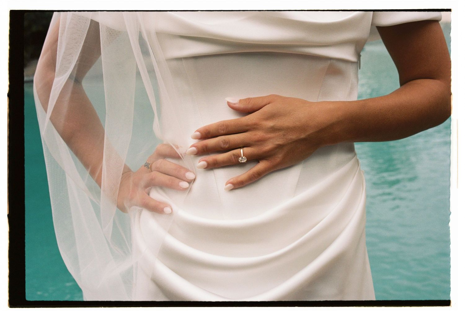 Close-up of a bride in a white satin gown with a veil, hands on waist, wearing a diamond engagement ring.