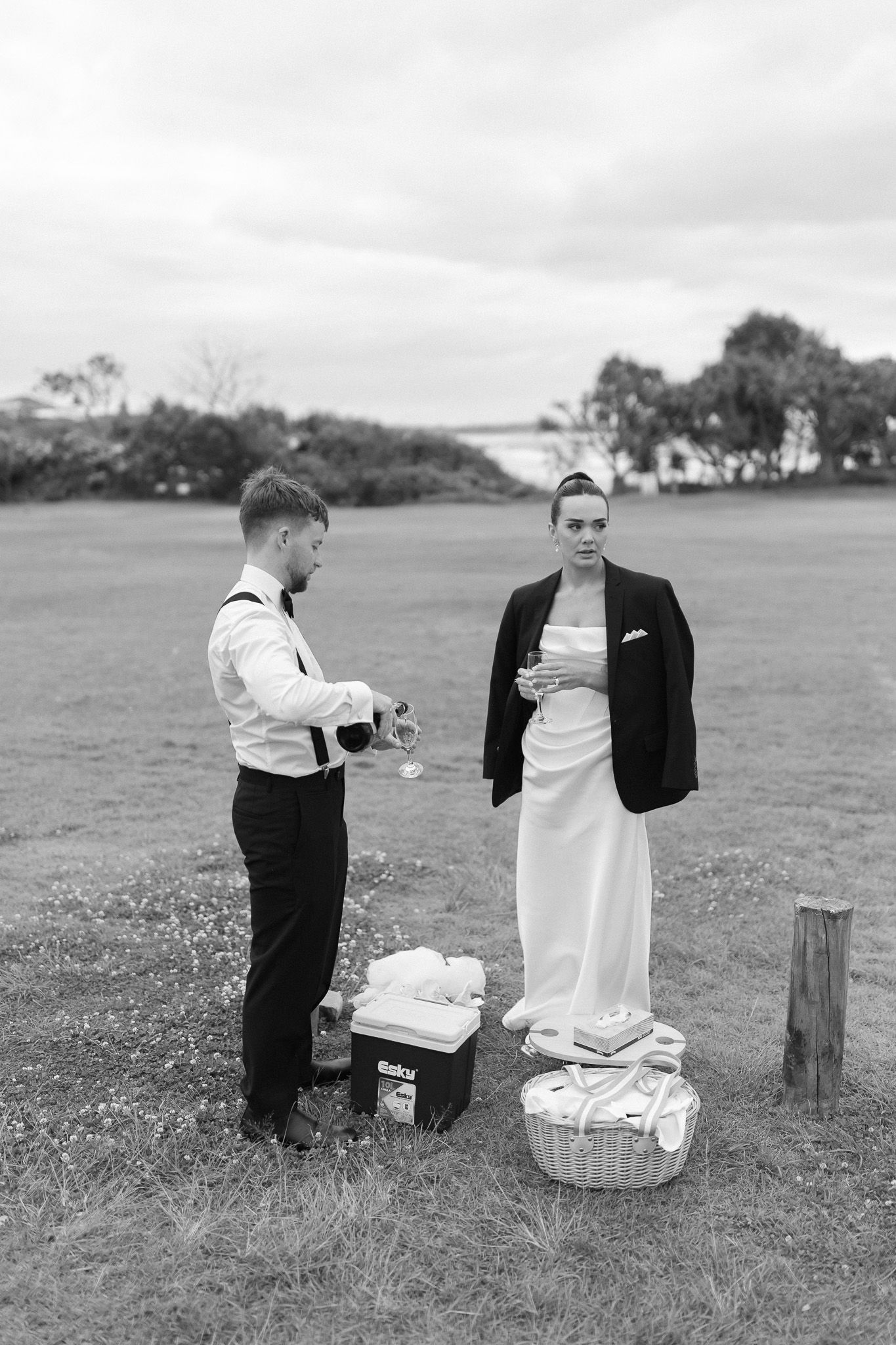 Black and white photo of bride and groom sharing champagne in open field at outdoor wedding.
