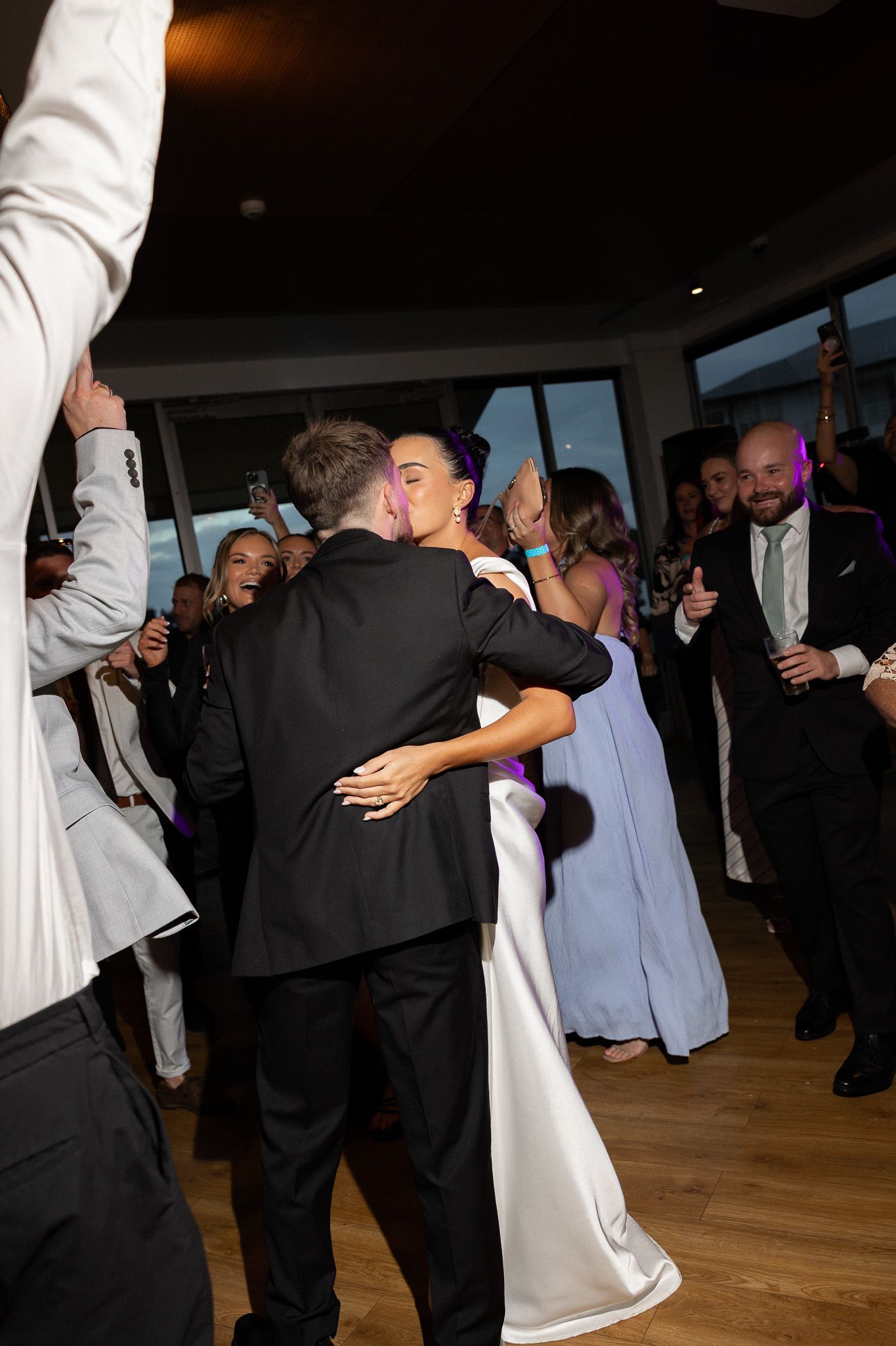 Bride and groom sharing first dance embrace surrounded by guests at wedding reception.