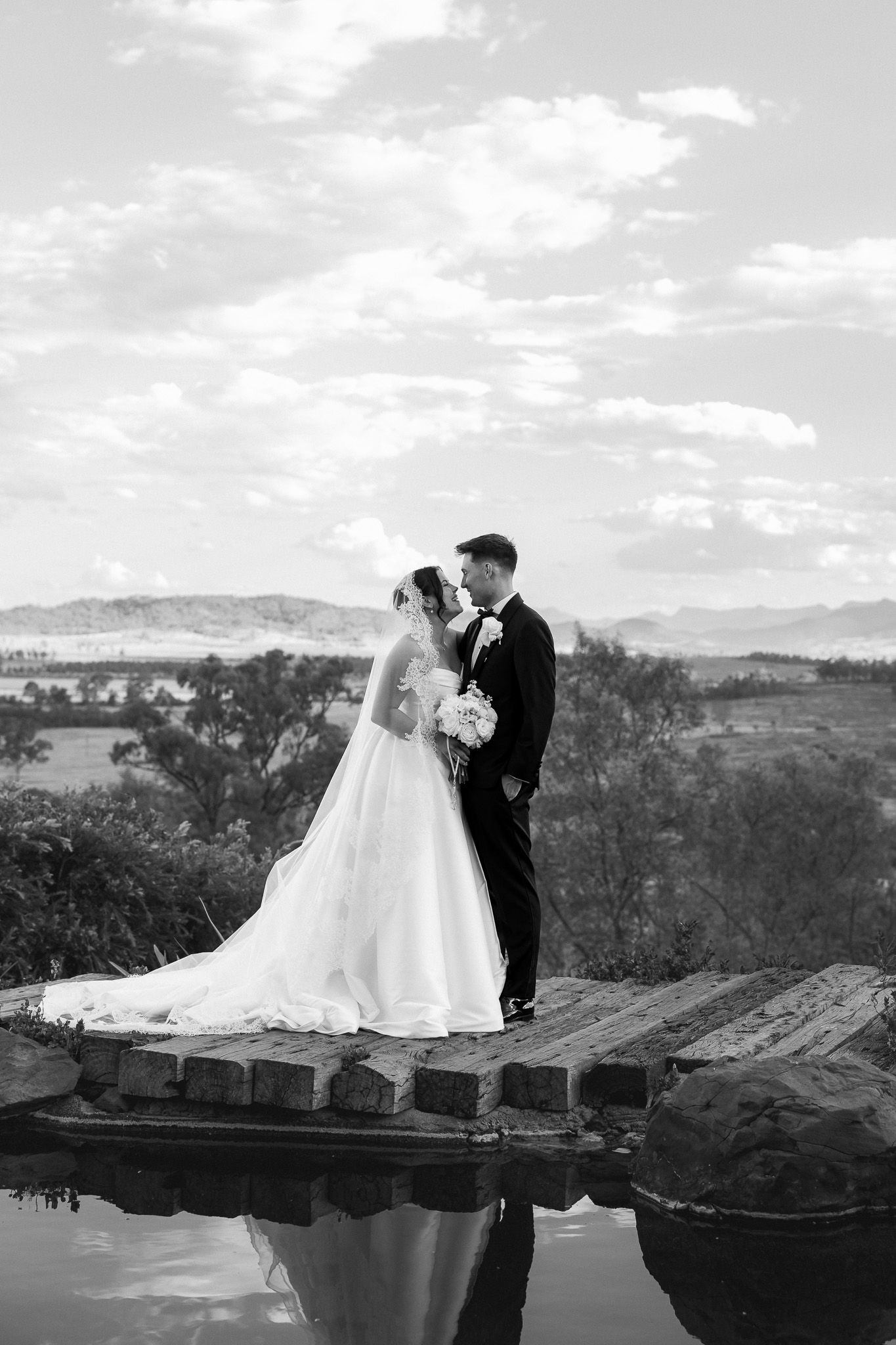 Black and white photo of bride and groom sharing a romantic moment on a dock with mountain landscape backdrop.