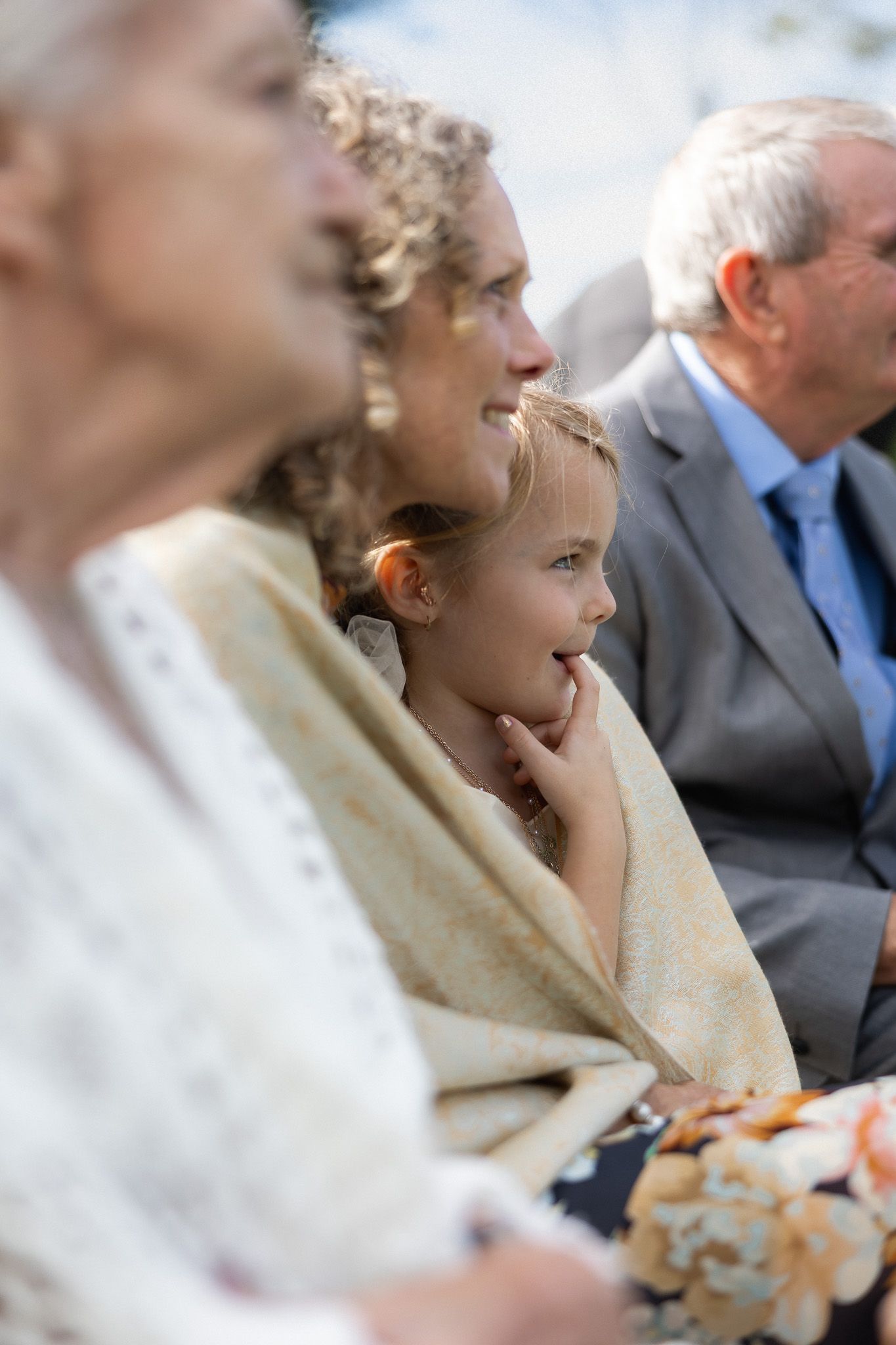 A young girl wrapped in a blanket sits with family members during an outdoor wedding ceremony.