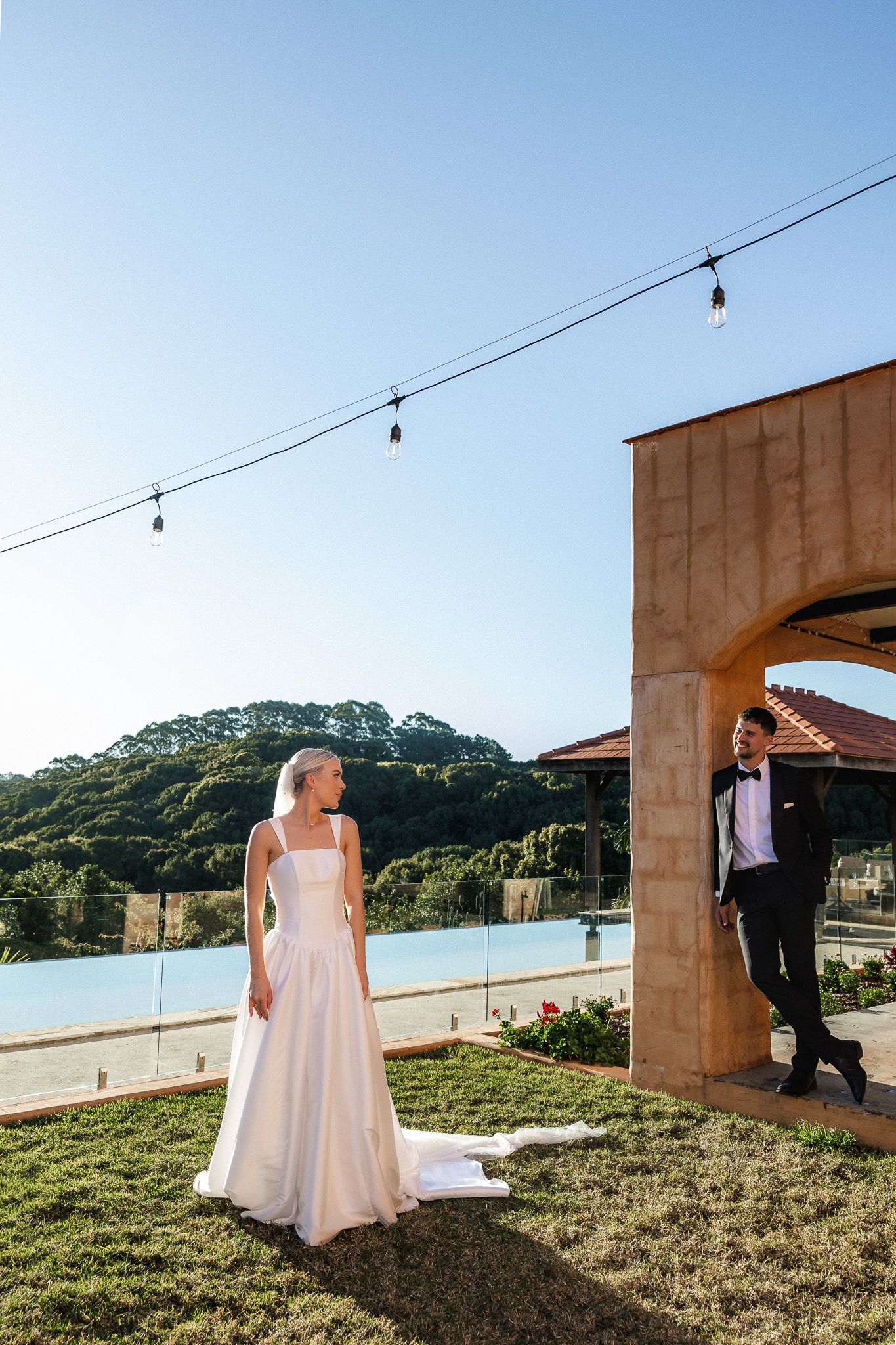 Bride and groom share a romantic moment outdoors near a pool with scenic hillside views.