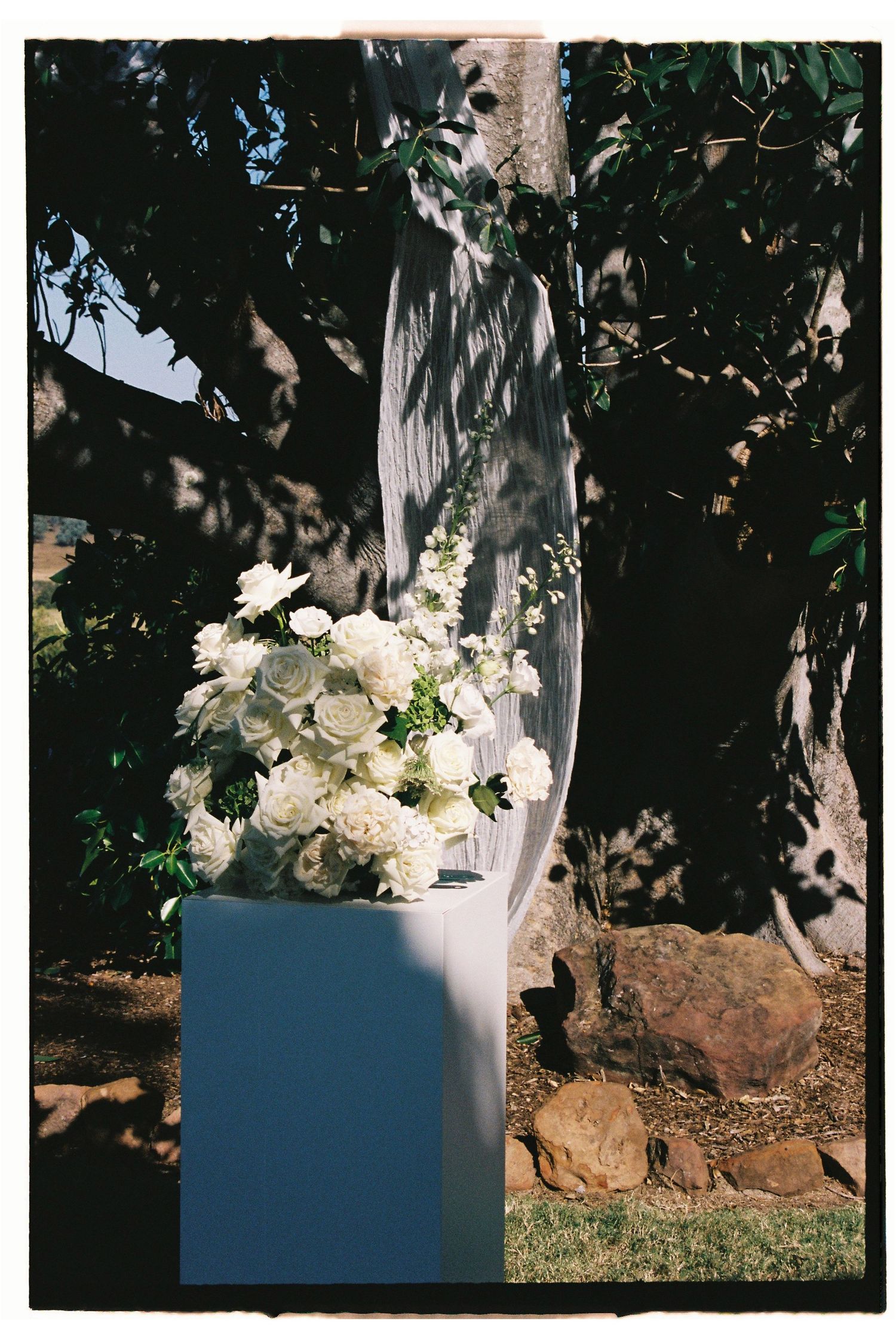 Elegant white floral arrangement on pedestal with draped fabric at outdoor wedding ceremony.