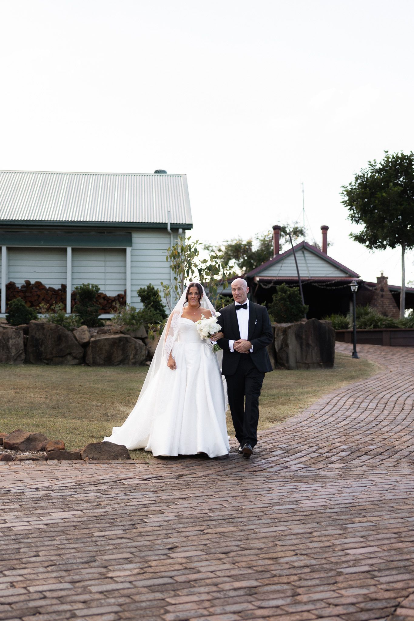 Bride in white gown and veil walks with escort on brick path toward wedding ceremony.