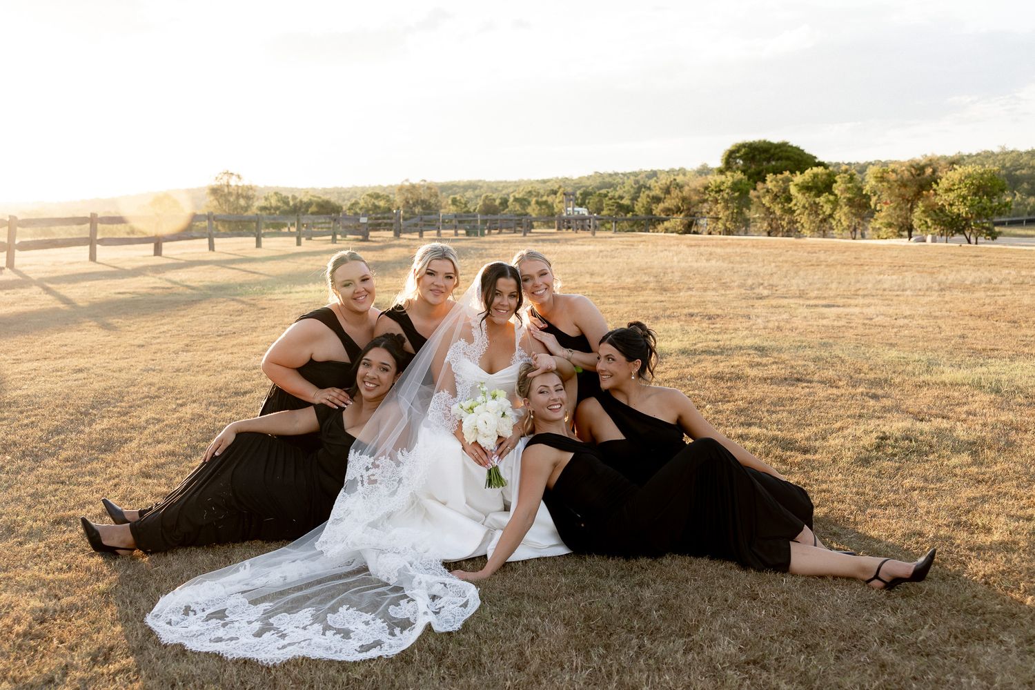 Bride in white gown with bridesmaids in black dresses posing together on golden grass field at sunset wedding.