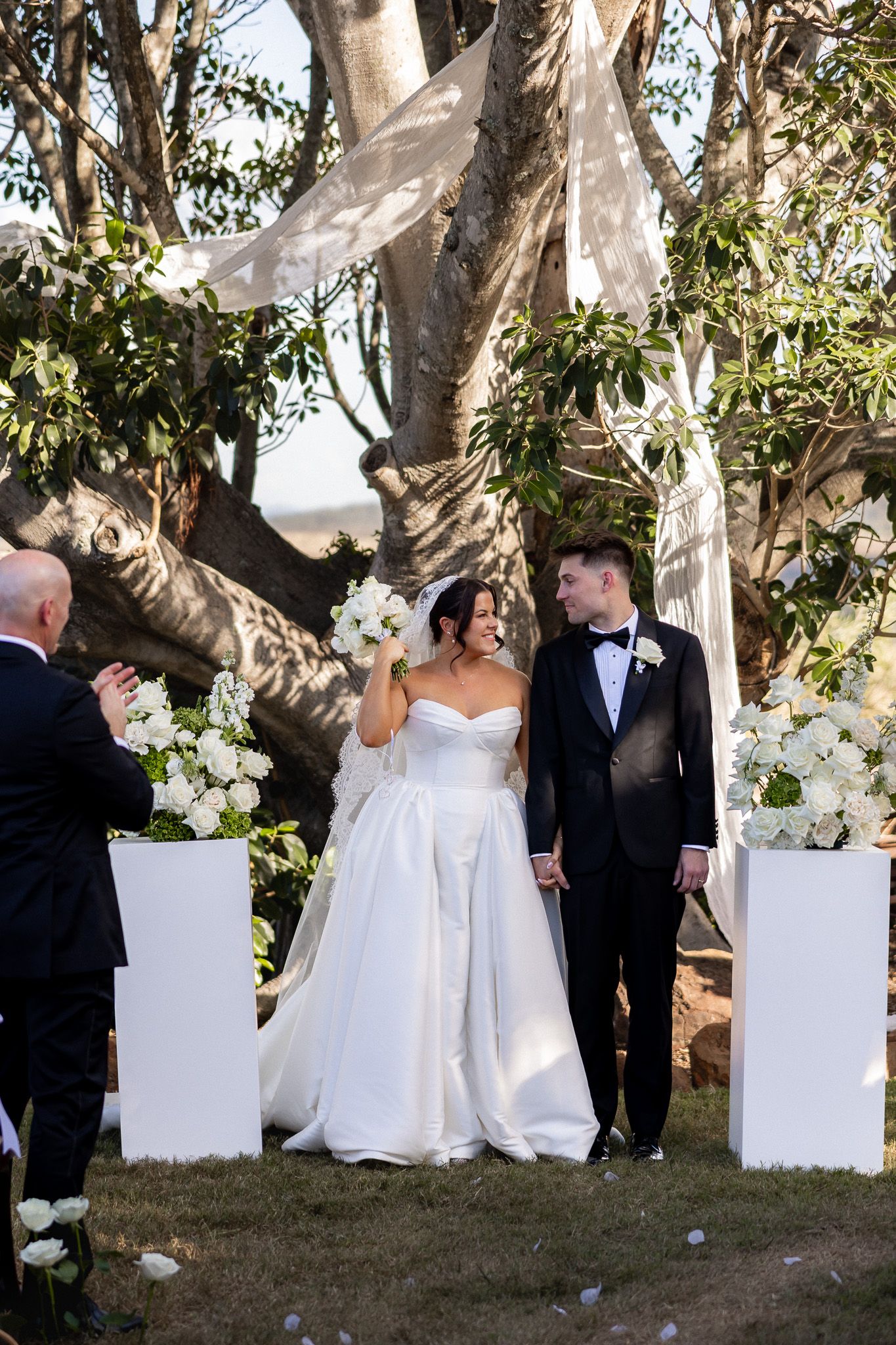 Newlyweds hold hands and smile at outdoor ceremony altar adorned with white flowers.