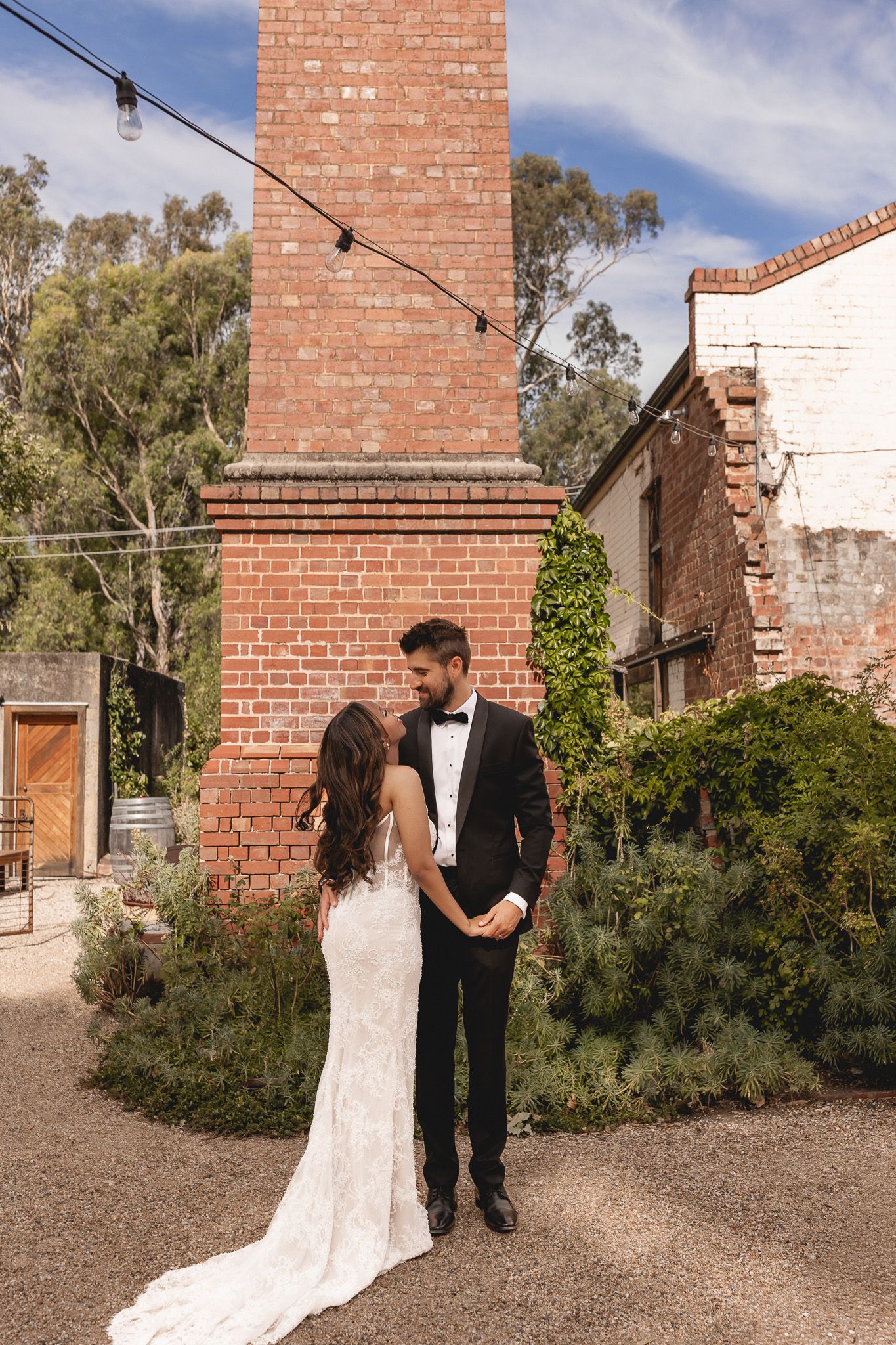 Bride and groom sharing a romantic moment near a rustic brick chimney at an outdoor wedding venue.