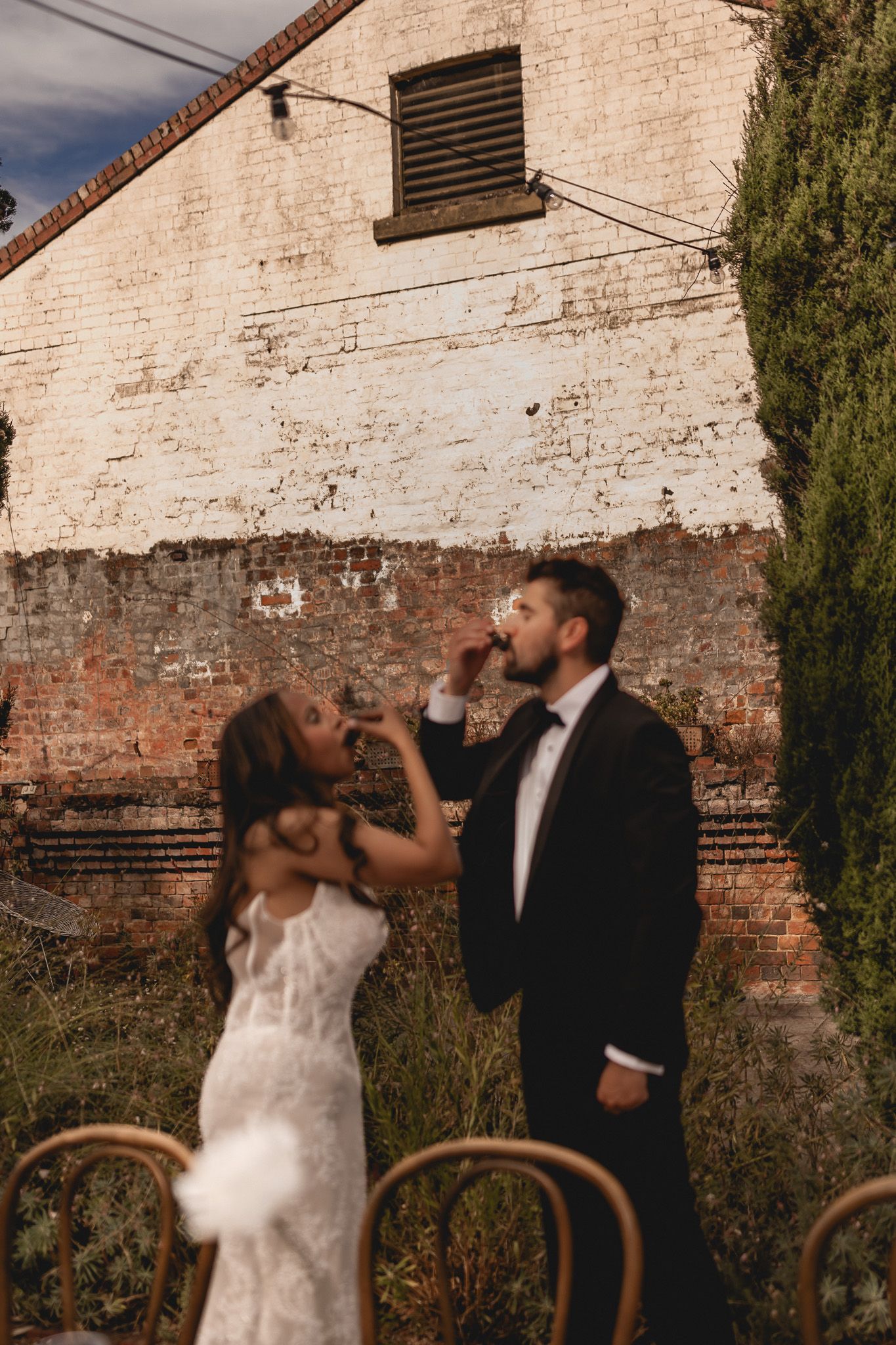 Bride feeds groom wedding cake outdoors against rustic brick wall, both dressed in elegant white gown and black tuxedo.