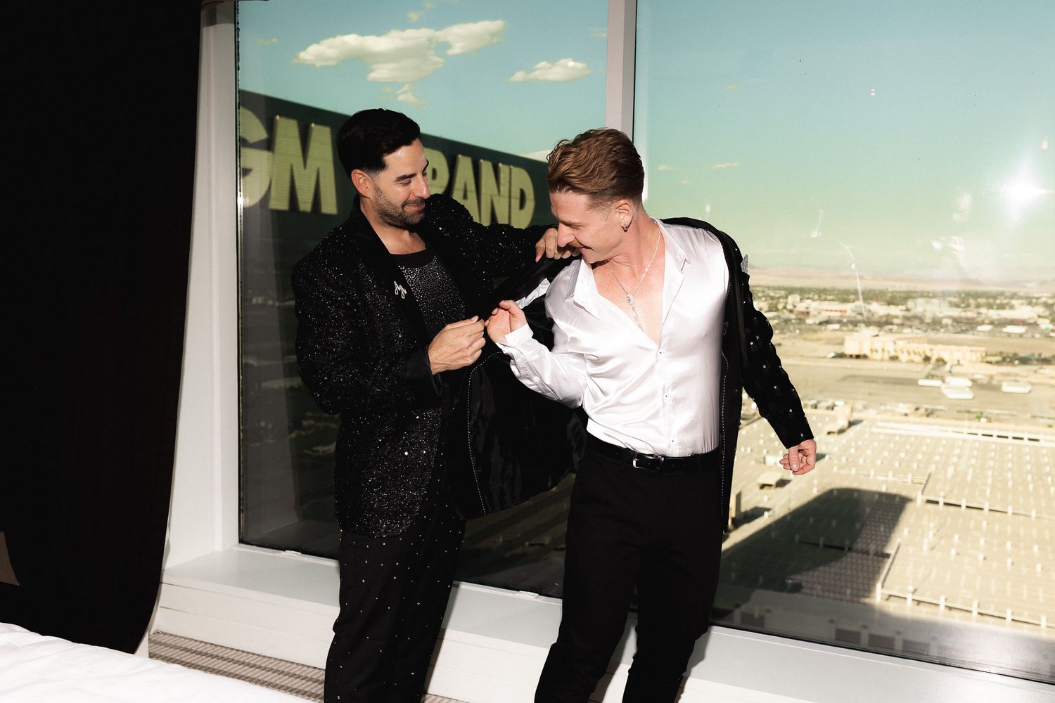 Two men in formal attire pose together near a high-rise window, one in sequins and one in a white shirt.