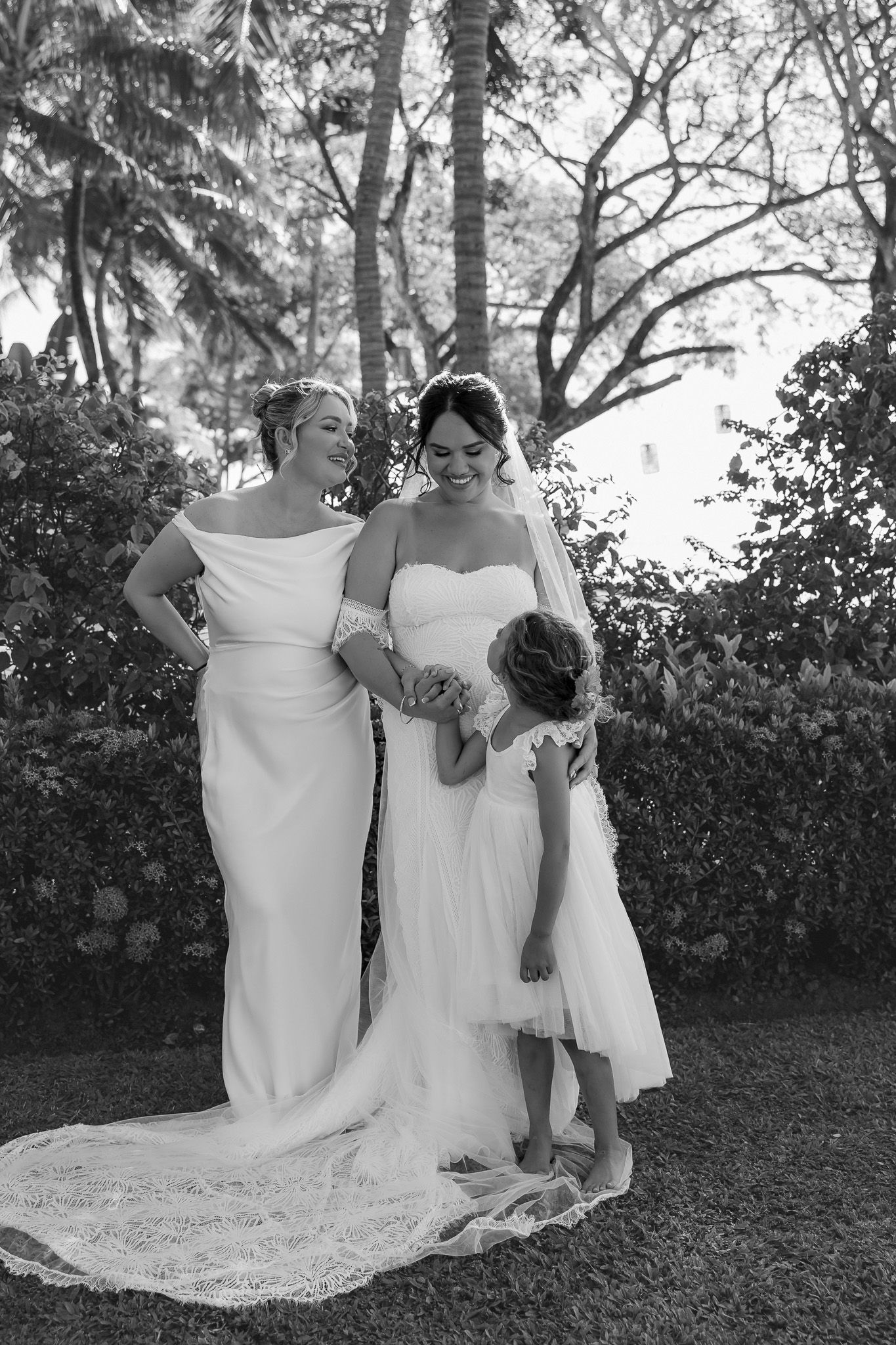 Black and white photo of two brides in white gowns smiling with flower girl outdoors.
