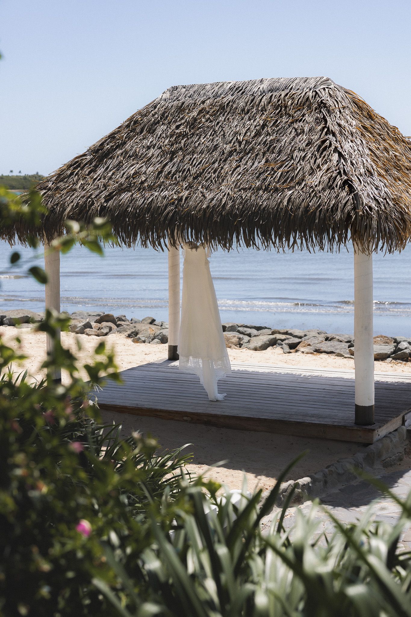 Thatched roof beach gazebo with white draping overlooks a serene tropical shoreline under clear blue skies.
