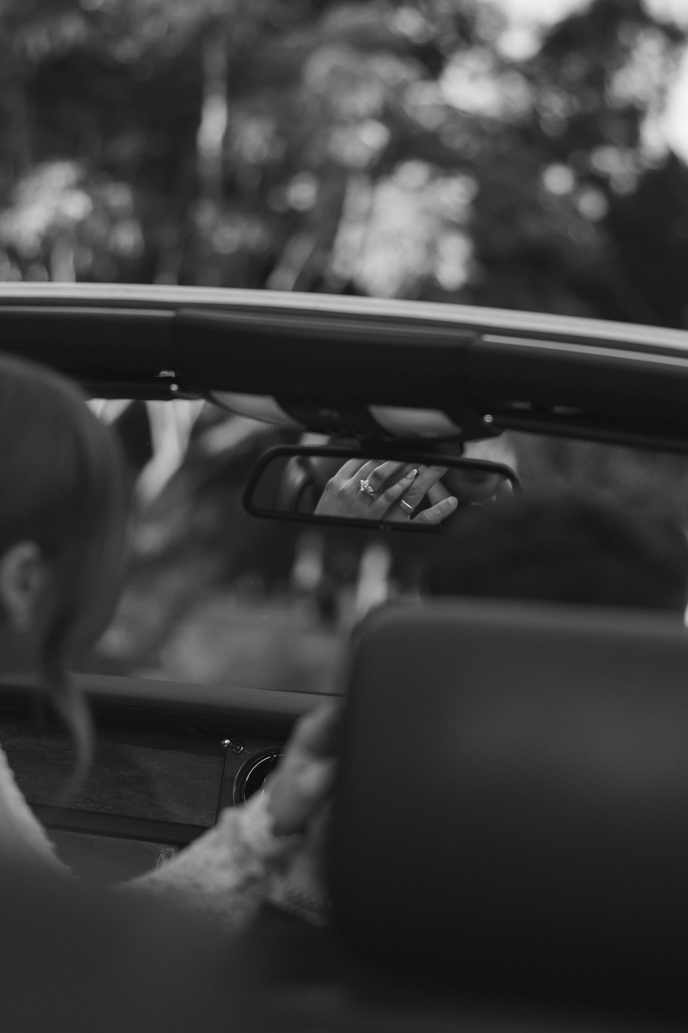 Black and white photo of a person driving a convertible car, reflected in the rearview mirror surrounded by trees.