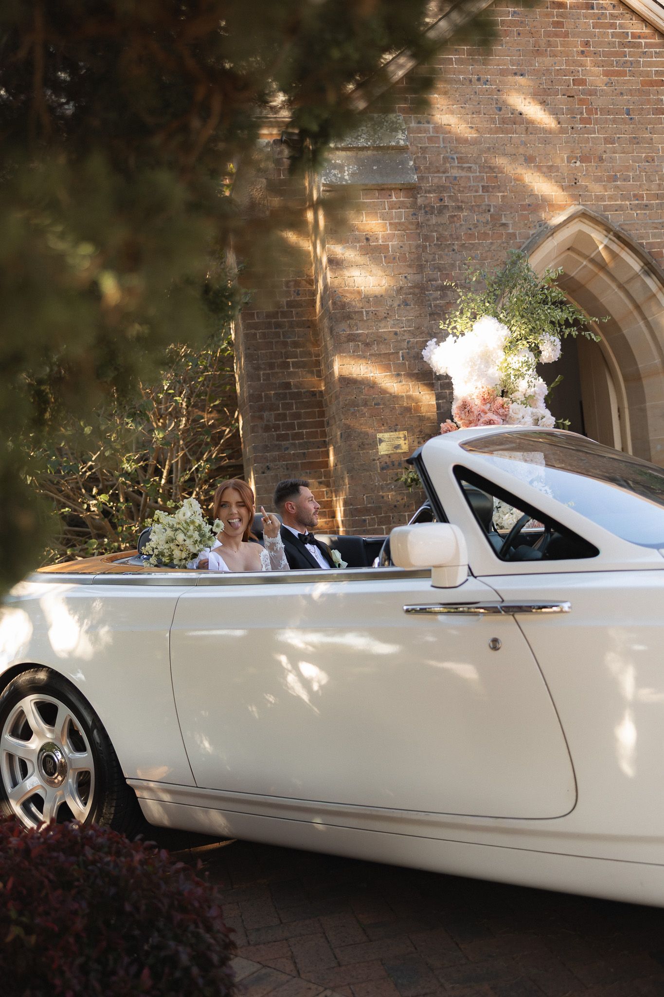 Bride and groom seated in a white Rolls-Royce convertible outside a brick church on their wedding day.