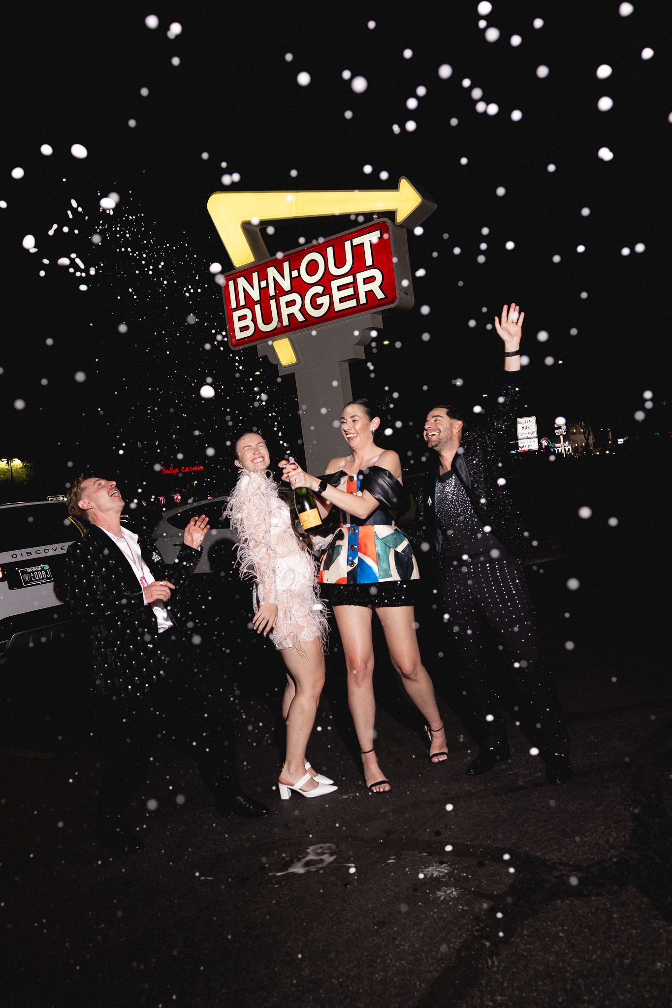 Group of friends celebrate with champagne and confetti in front of an In-N-Out Burger sign at night.