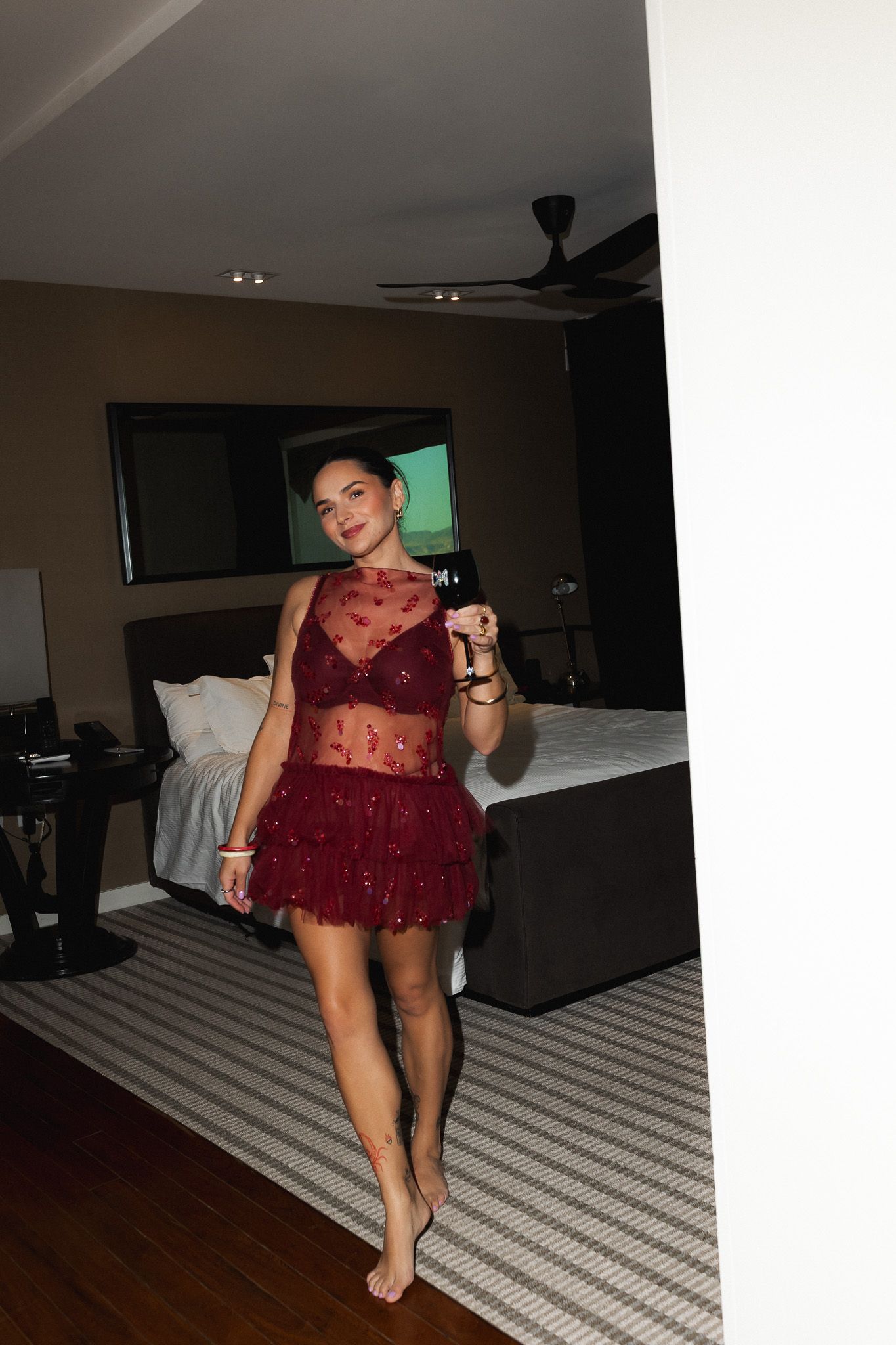 Woman in red lace mini dress holding a wine glass, standing barefoot in a modern bedroom.