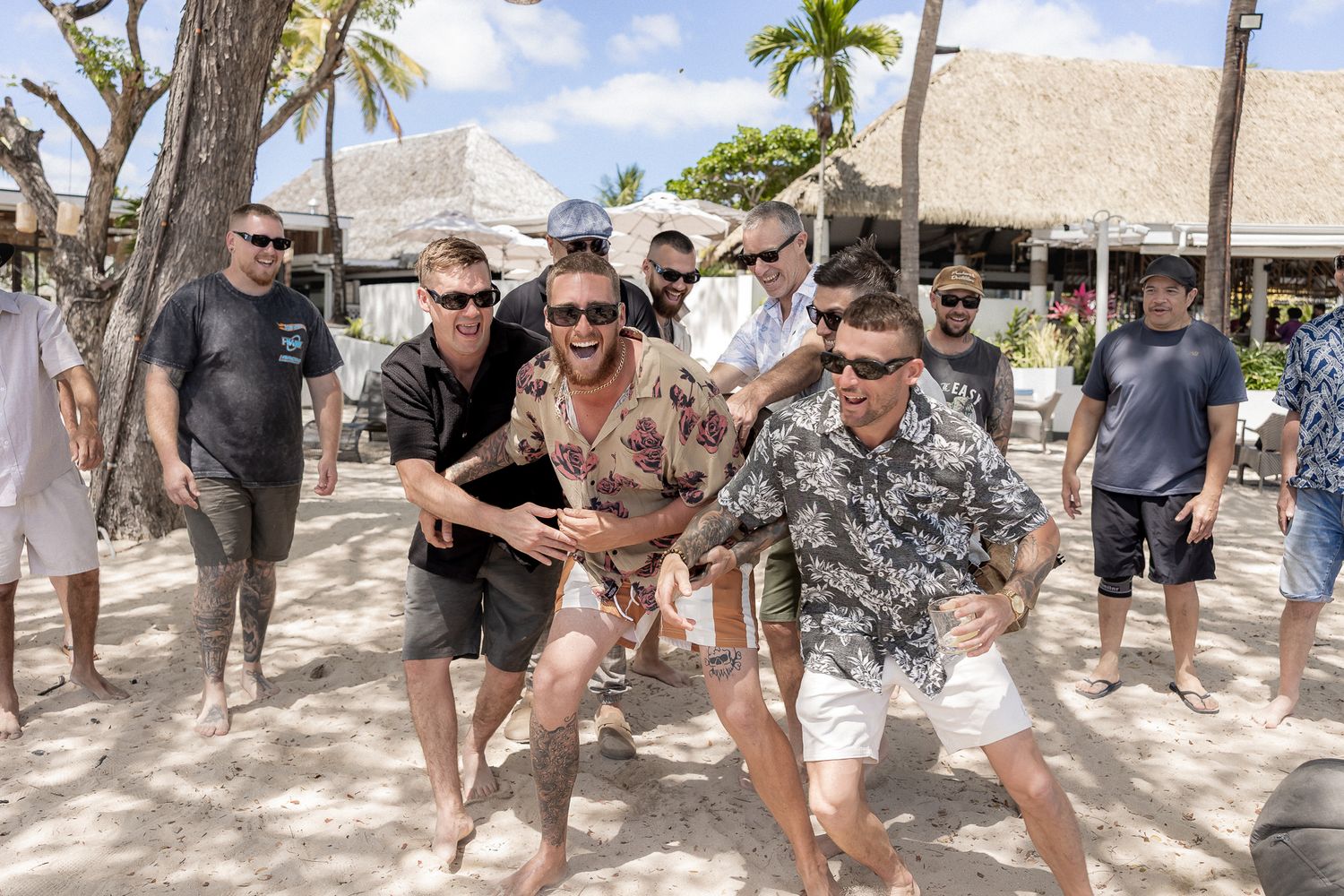 Group of men posing and dancing on a tropical beach resort, wearing casual summer clothes on a sunny day.