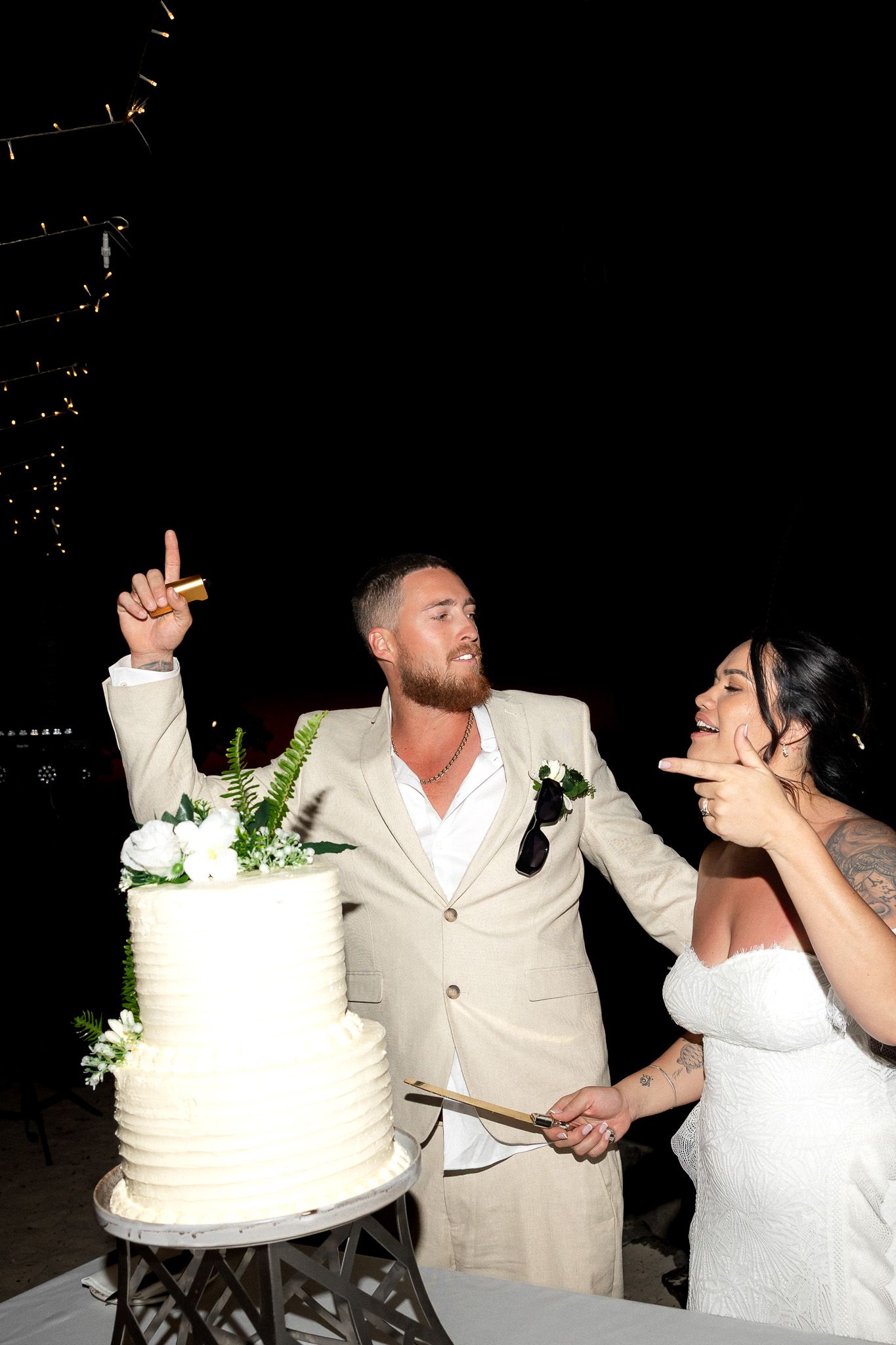 Bride and groom laugh together while cutting their white two-tier wedding cake at a nighttime reception.