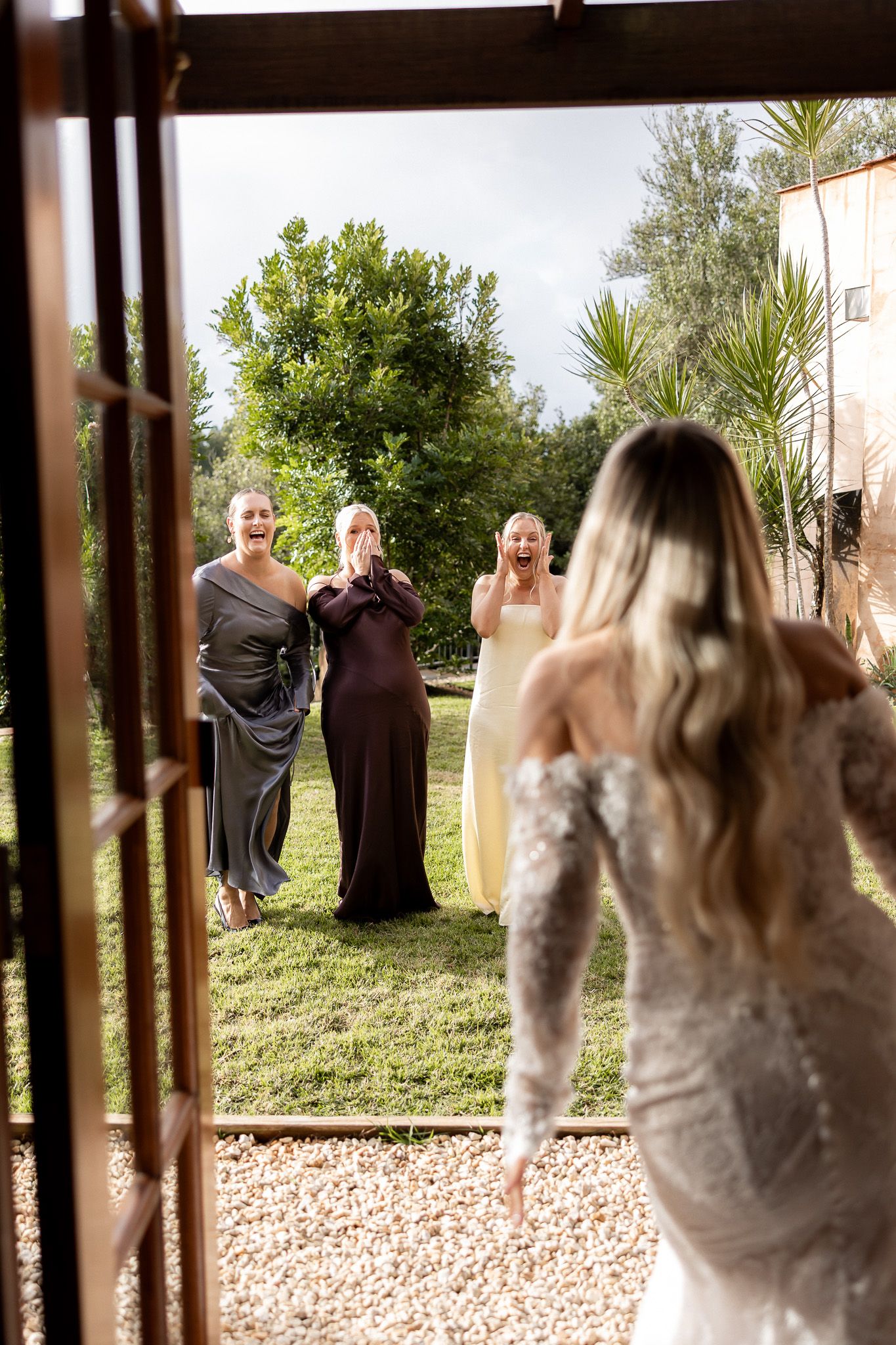Bride in lace gown reveals herself to emotional bridesmaids in colorful dresses outdoors at a garden wedding venue.