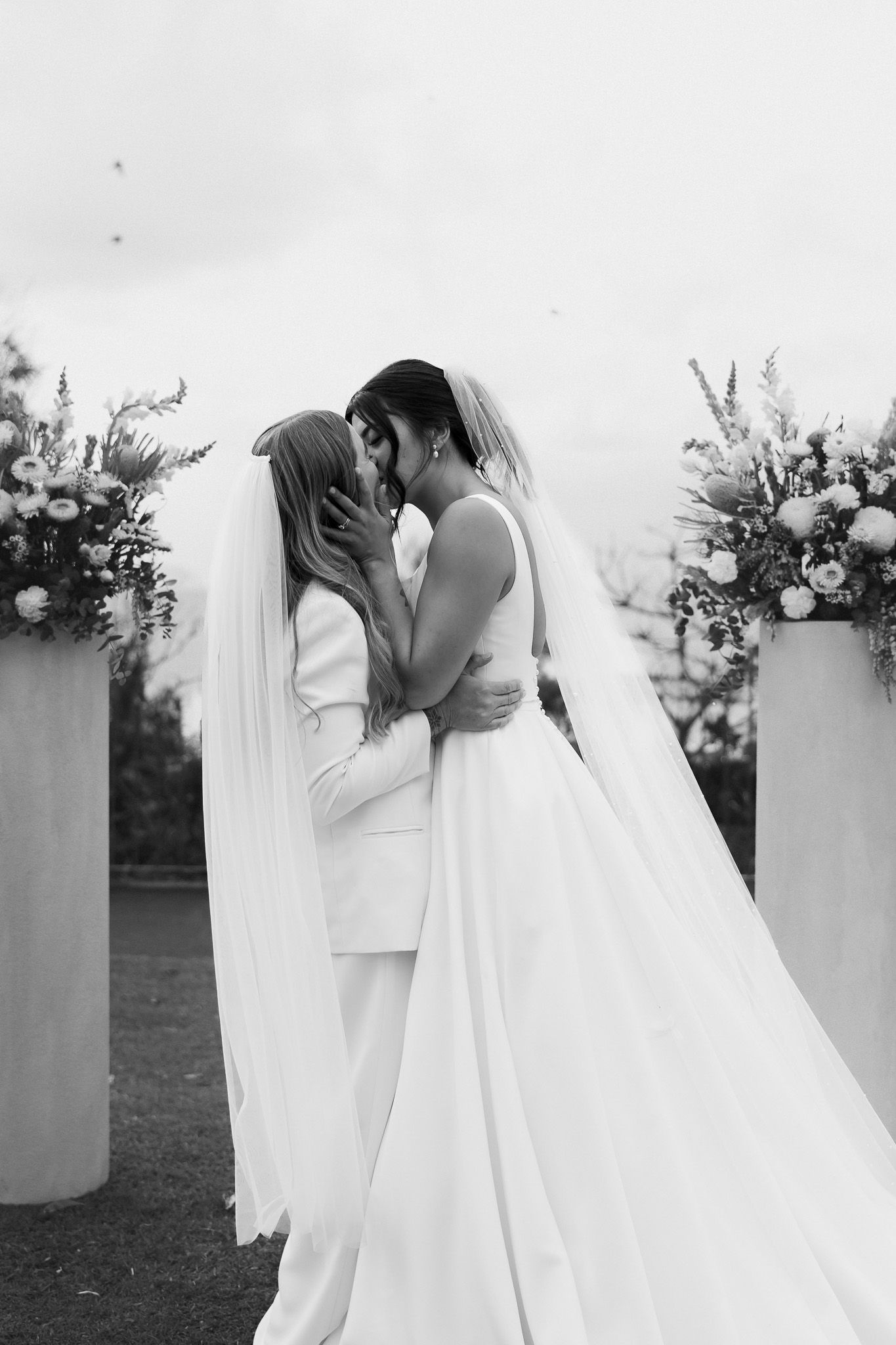 Black and white photo of two brides sharing a kiss at outdoor wedding ceremony.