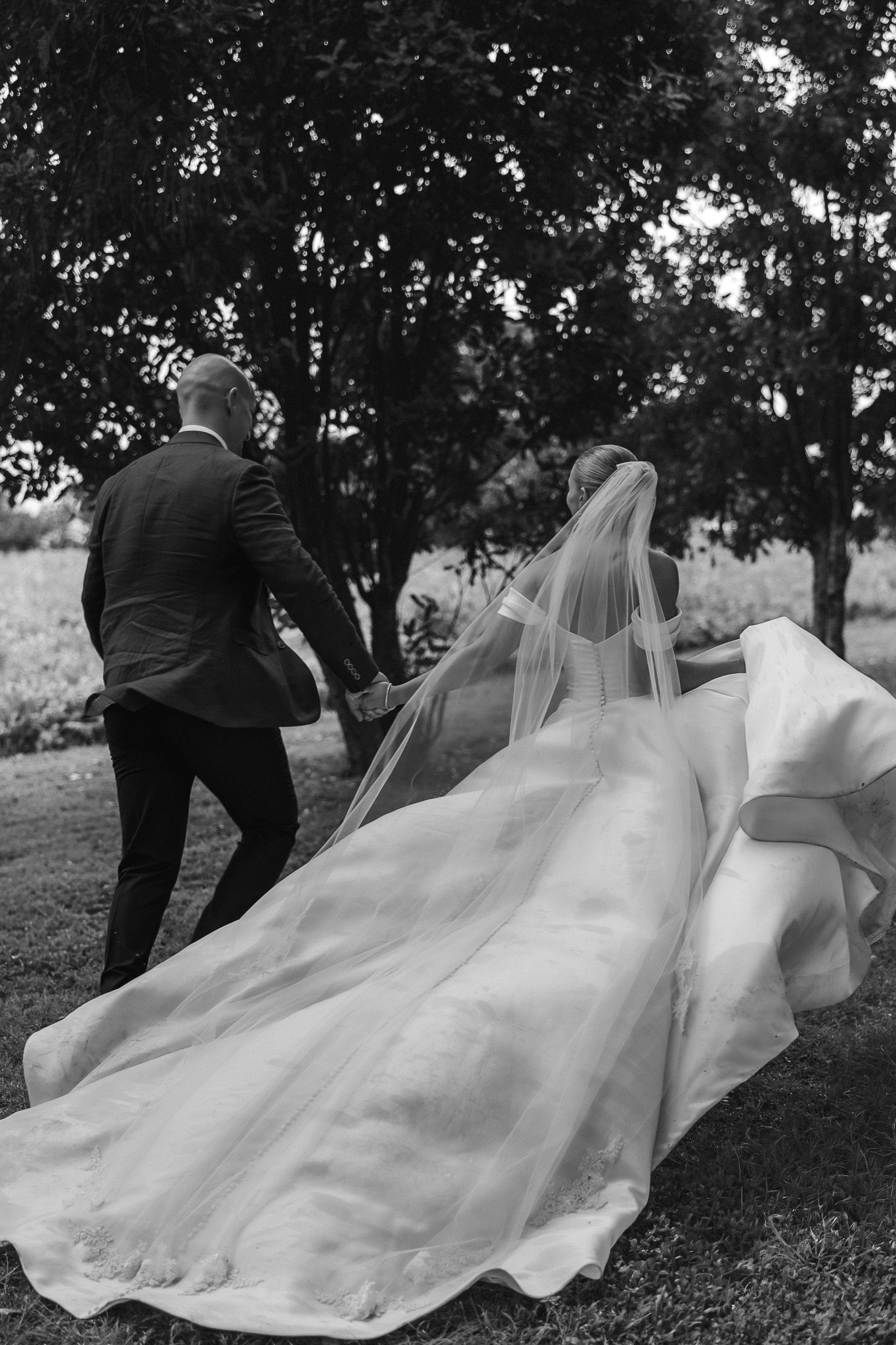 Black and white photo of bride and groom walking hand in hand, bridal veil flowing behind them.