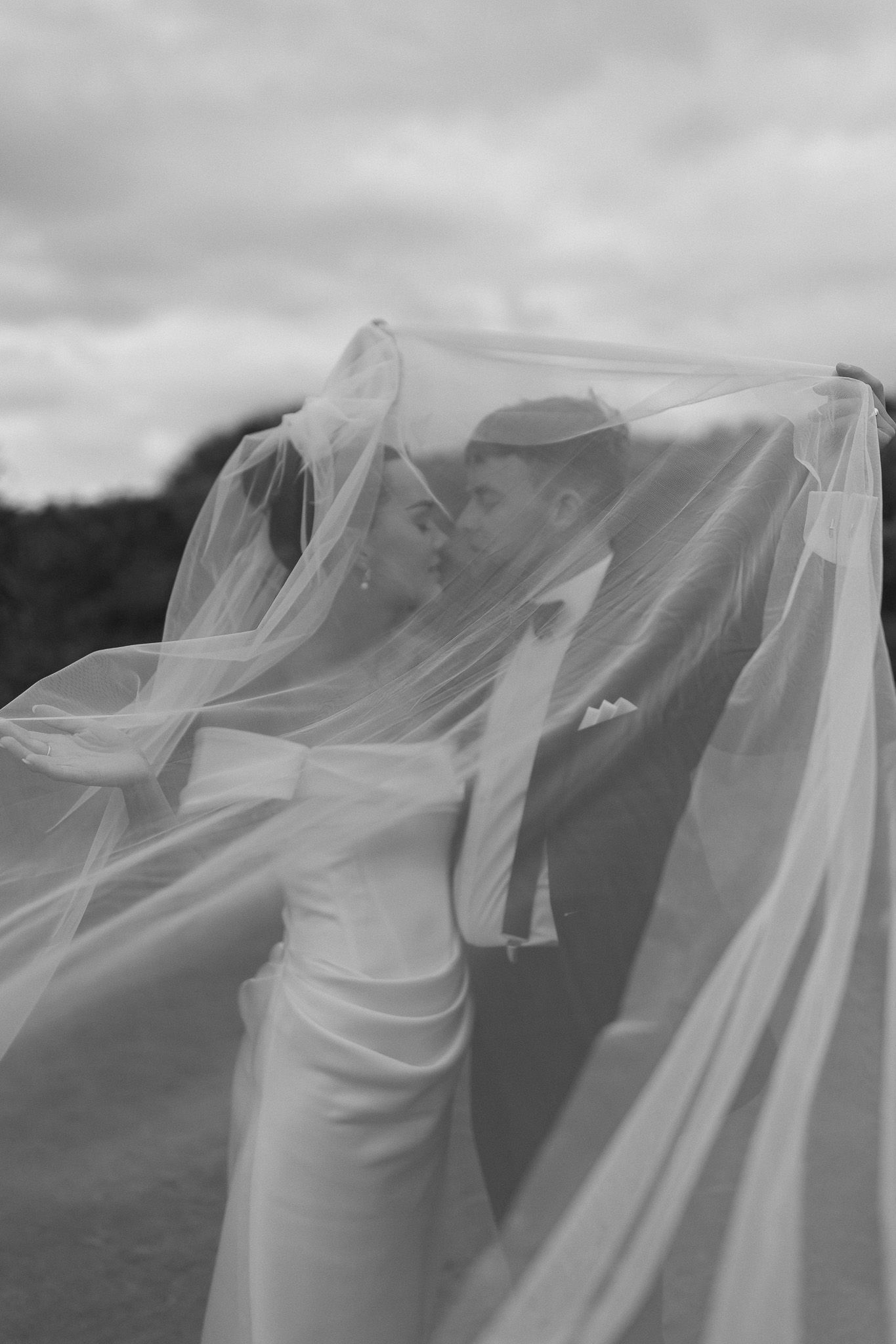 Black and white photo of bride and groom kissing beneath flowing wedding veil.