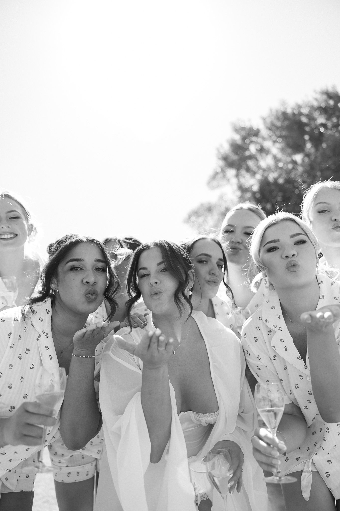 Black and white photo of bride and bridesmaids in robes blowing kisses, holding champagne glasses outdoors.
